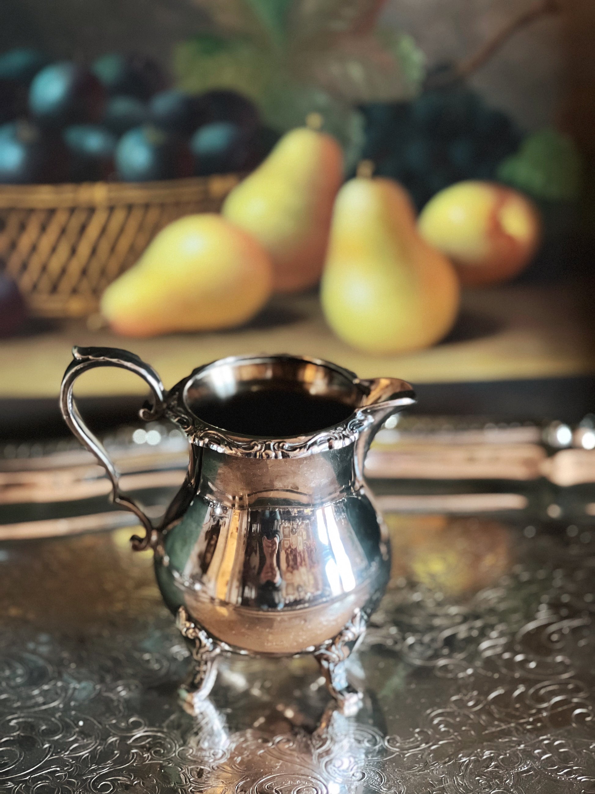 Silver creamer on a reflective surface with pears and plums in the background