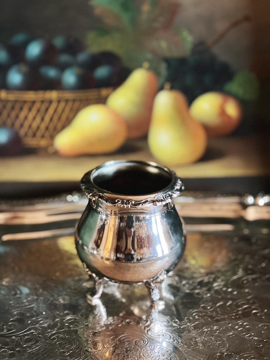 Silver container on a reflective surface with pears and plums in the background