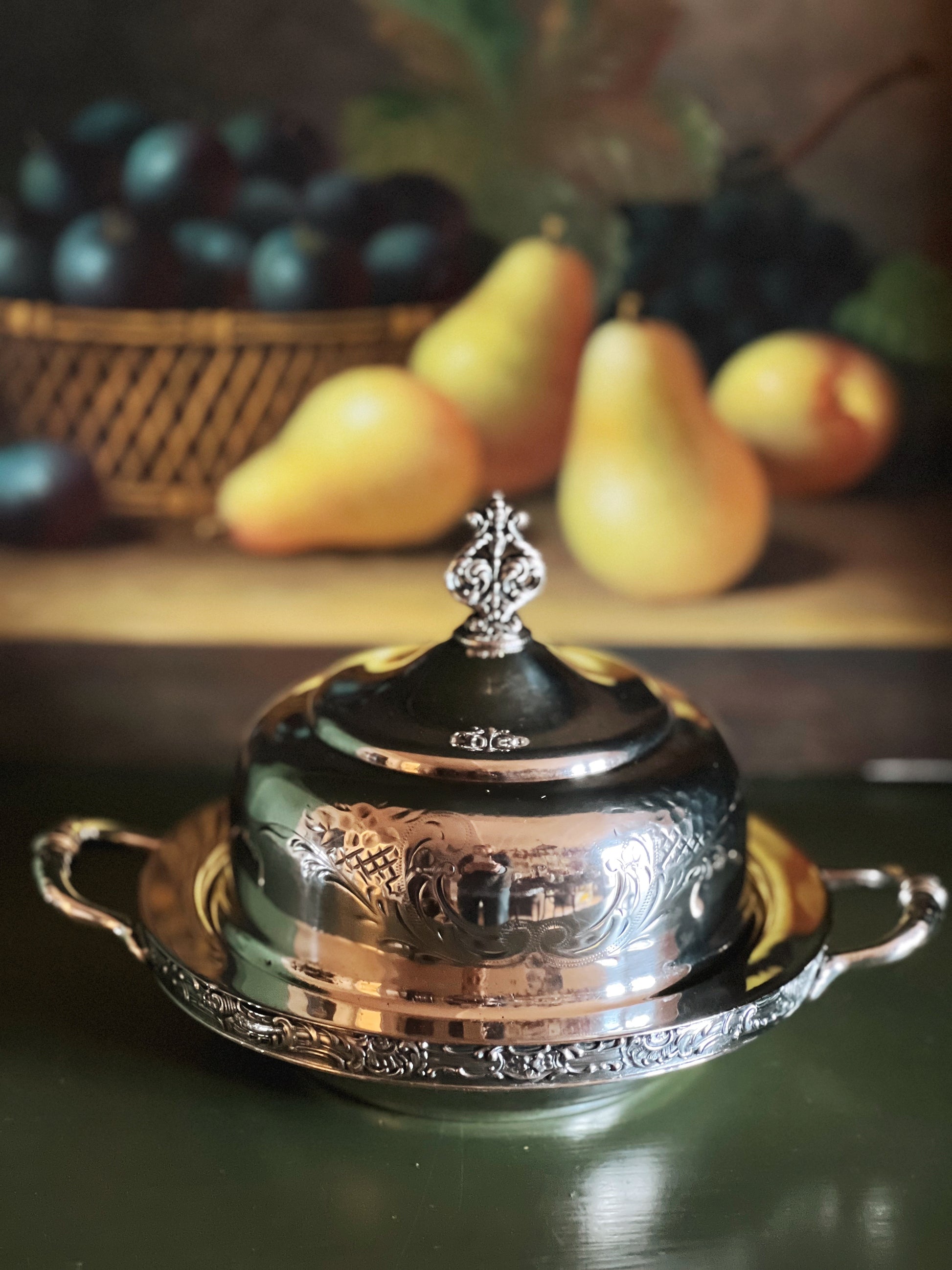 Decorative silver dish with a lid on a table with fruits in the background