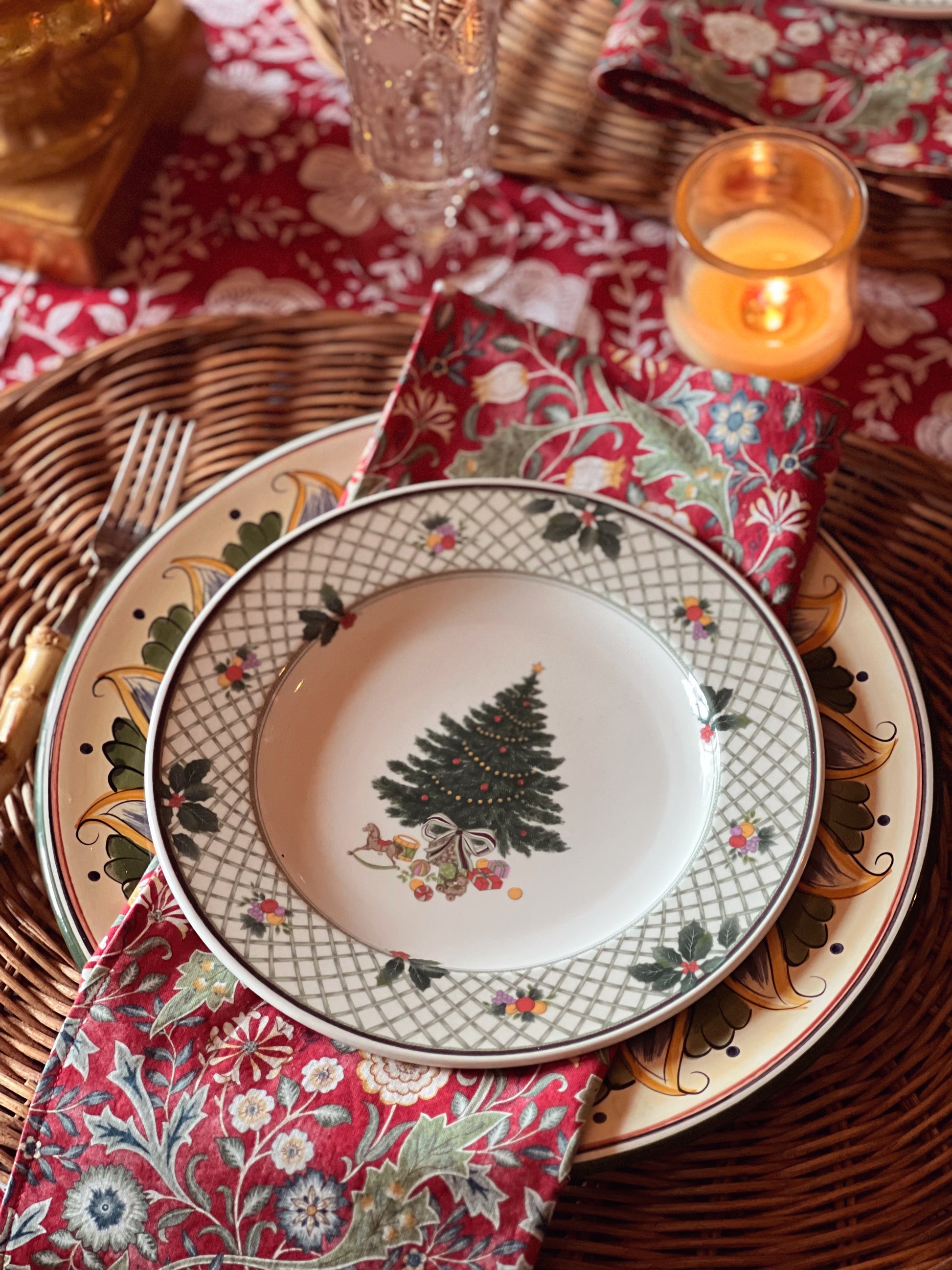 Decorative plate with Christmas tree design on a patterned tablecloth with candles and wicker basket.