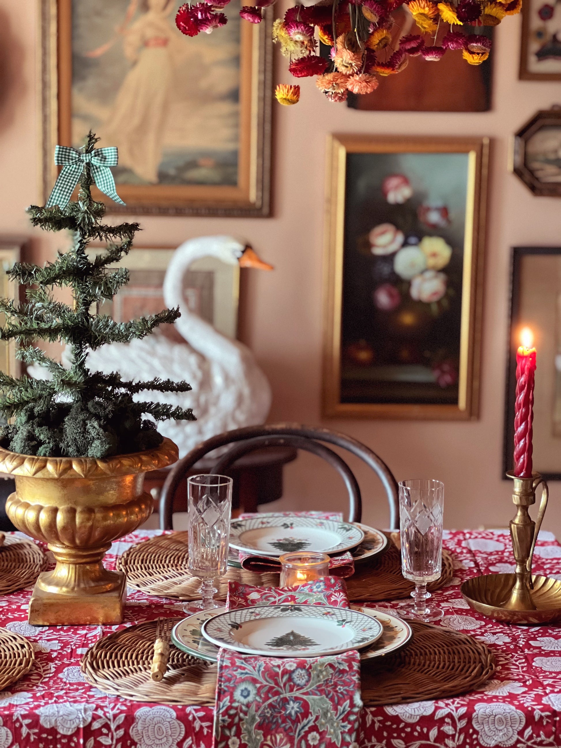 Decorative table setting with a small Christmas tree, candles, and patterned tablecloth in a room with framed artwork on the walls.