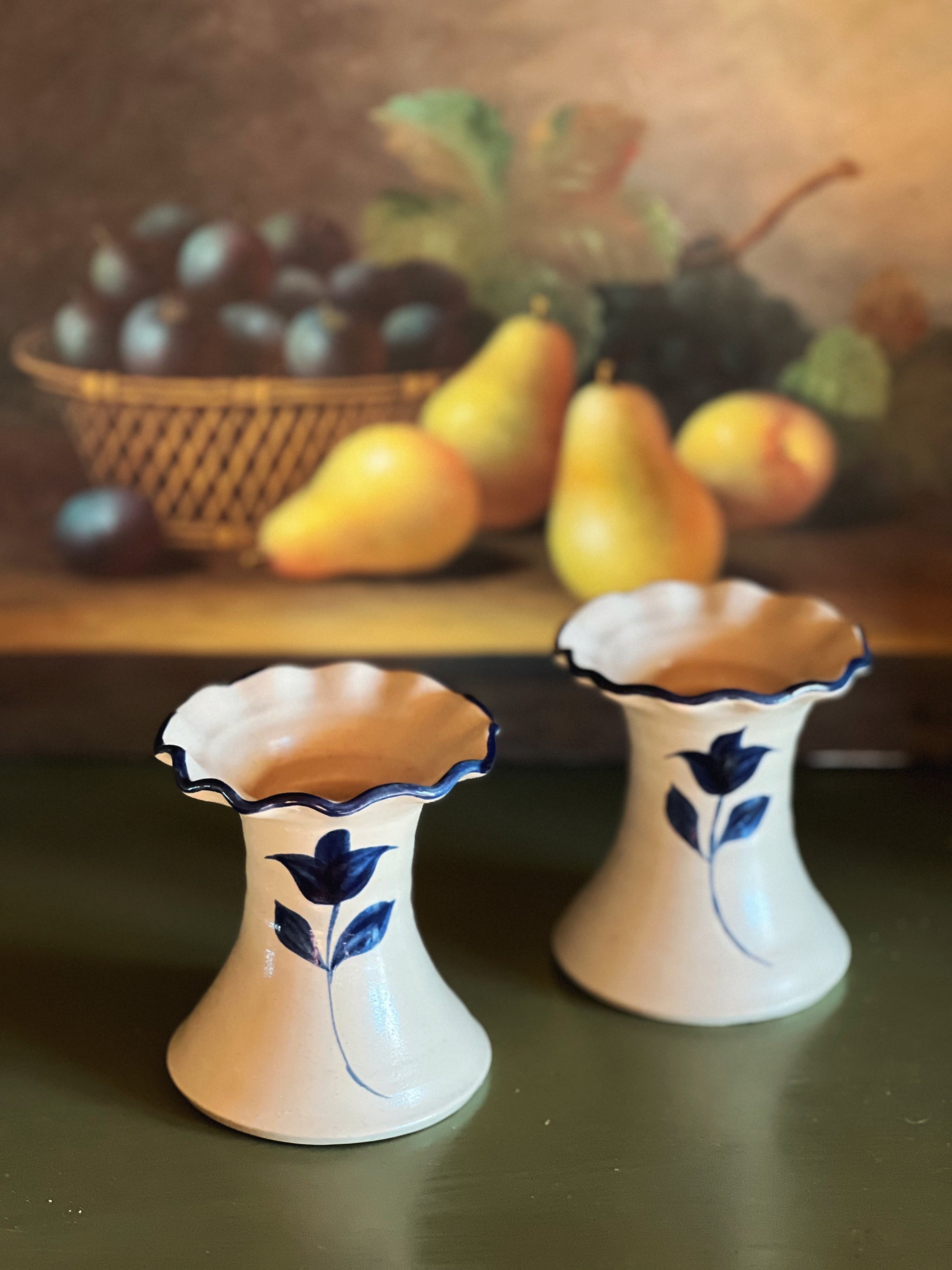 Two ceramic vases with floral designs on a table with a fruit basket in the background.
