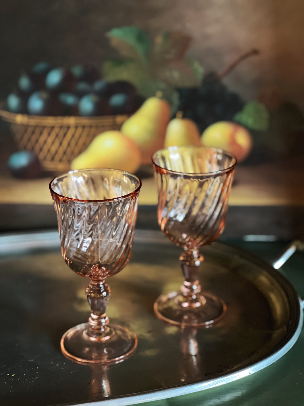 Two pink glass goblets on a reflective surface with a blurred background of fruits.