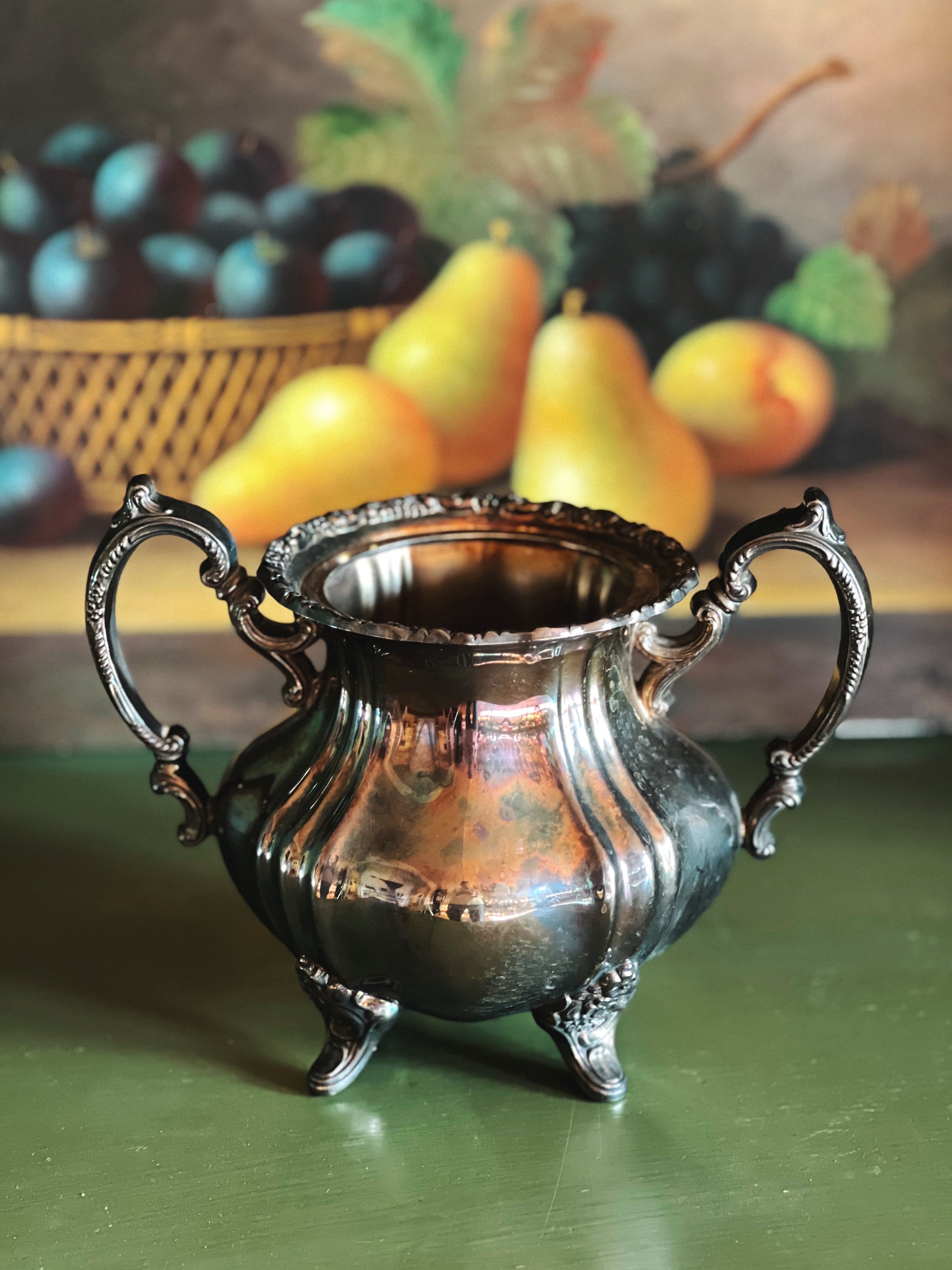 Silver ornate container on a green surface with fruit in the background