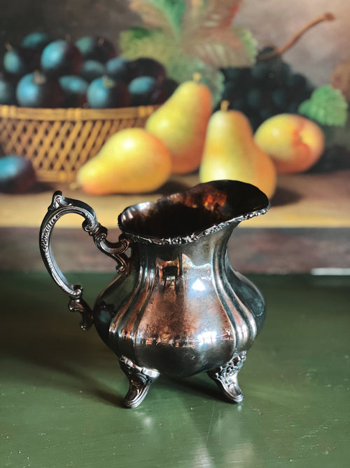 Silver pitcher on a green surface with fruit in the background