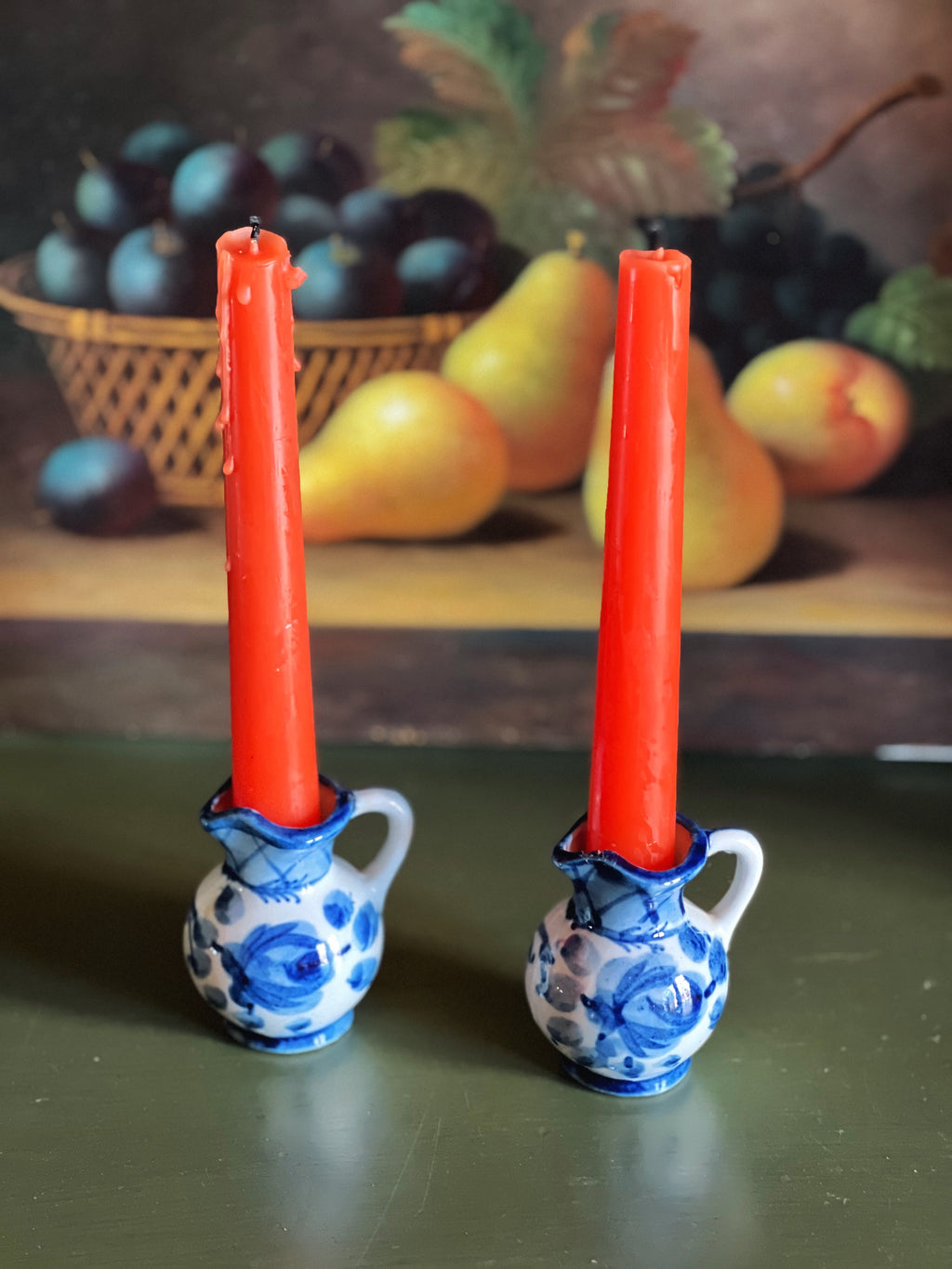 Two red candles in blue and white ceramic holders with a fruit basket in the background.