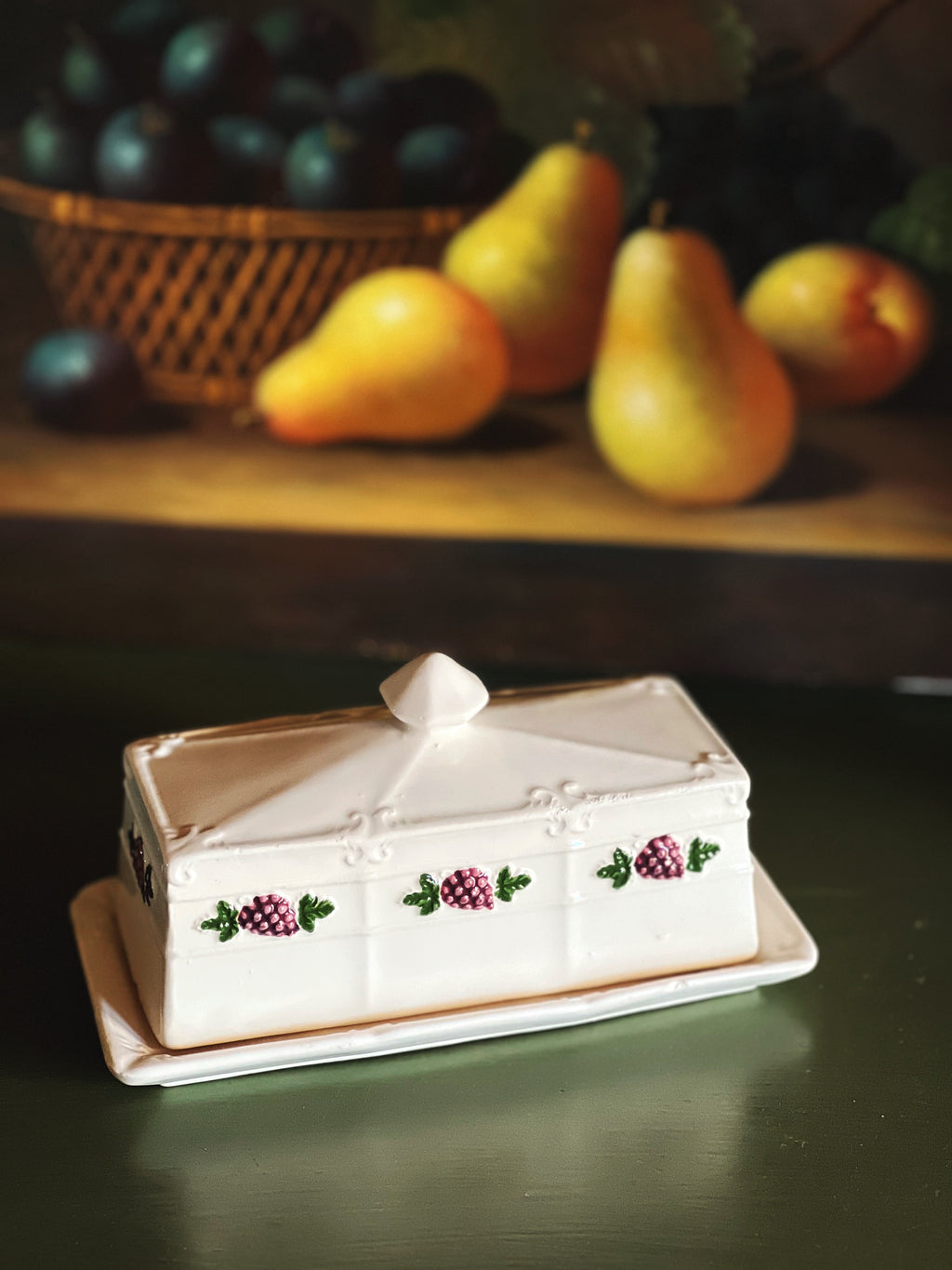 Decorative butter dish with grape design on a wooden surface with pears and plums in the background.