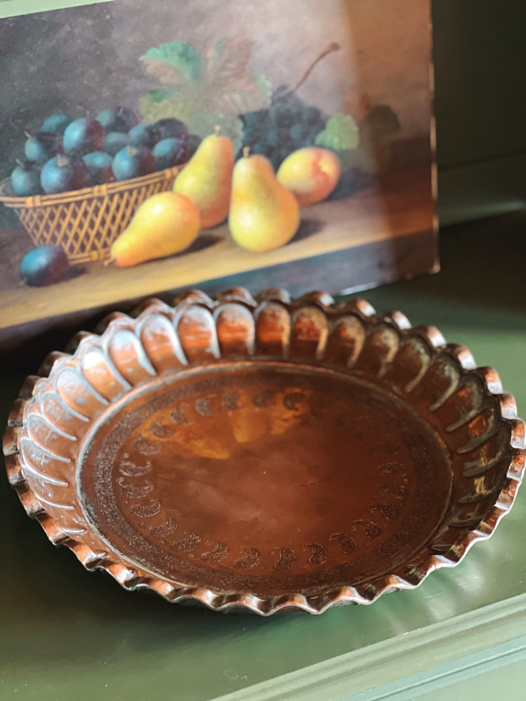 Copper tray with embossed design on a green surface, with a still life painting of fruit in the background.
