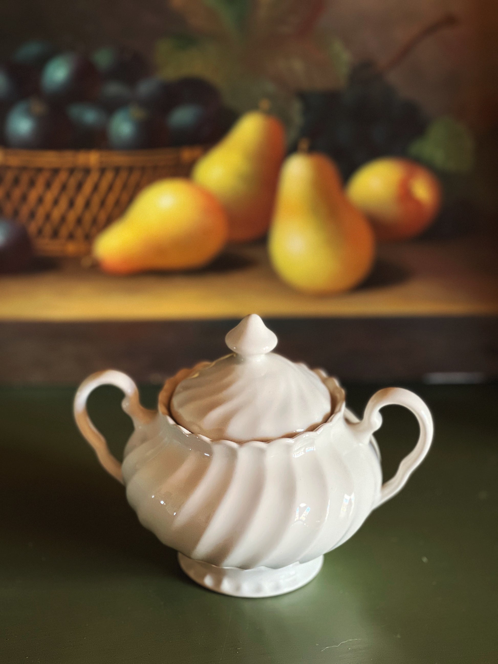 White ceramic sugar bowl with lid on a dark surface, with pears and plums in the background.
