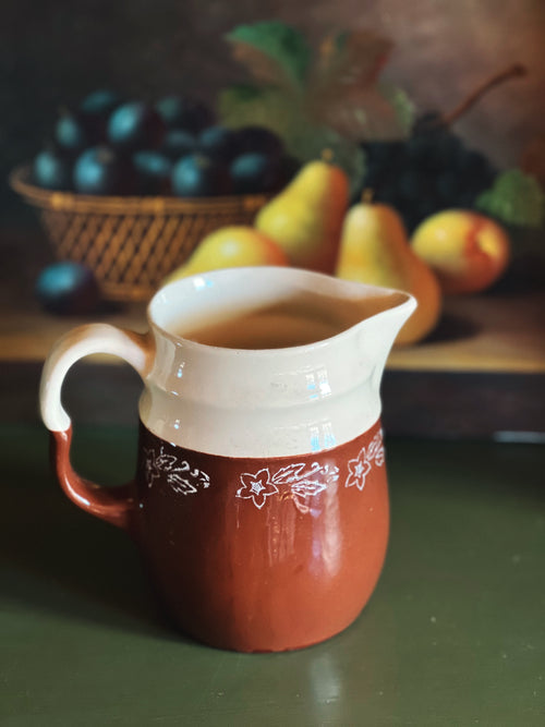 Red ceramic pitcher with white handle on a green surface, with fruits in the background.