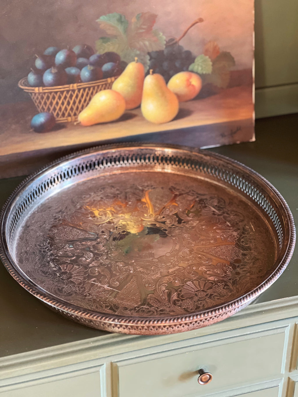 Decorative metal tray on a surface with a painting of fruit in the background