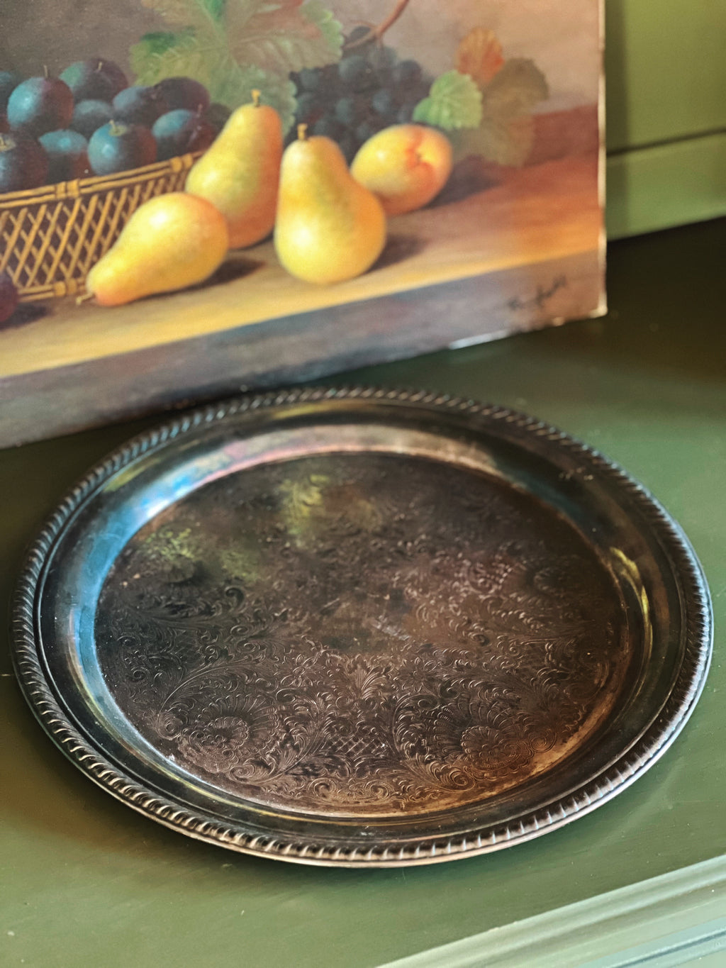 Vintage silver tray on a green surface with a painting of fruit in the background