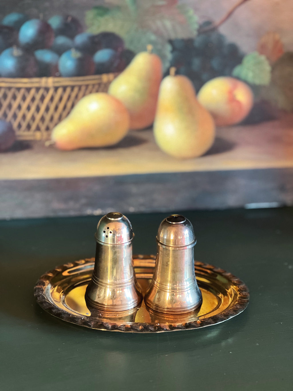 Two silver salt shakers on a decorative tray with a fruit painting in the background.