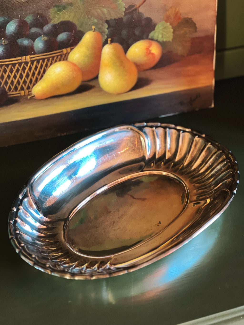 Silver dish on a reflective surface with a painting of fruit in the background