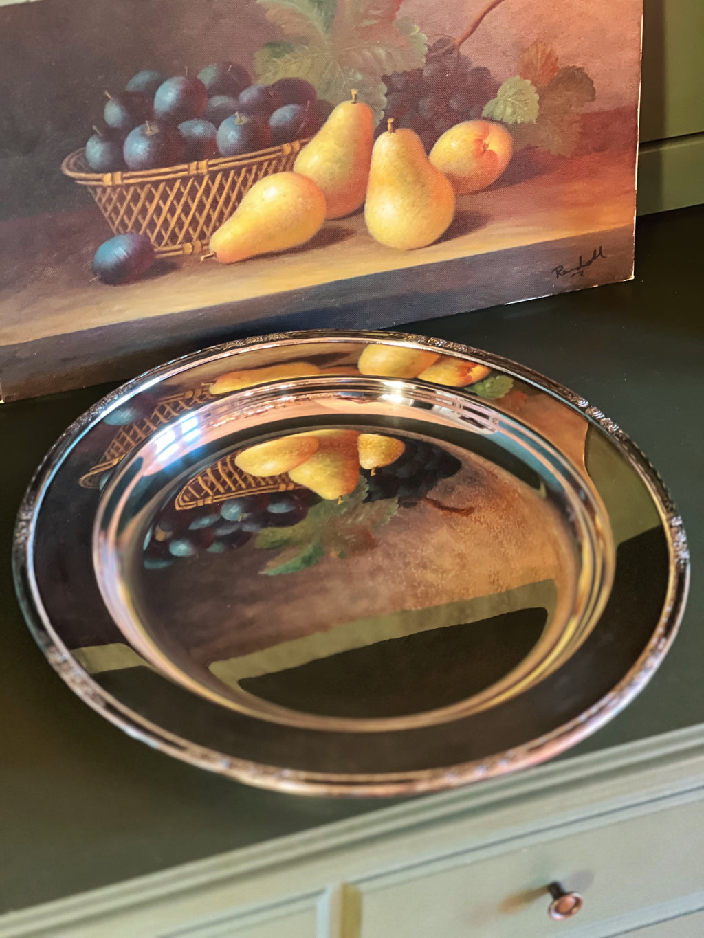 Decorative metal tray with fruit design on a green surface, below a painting of pears and grapes.