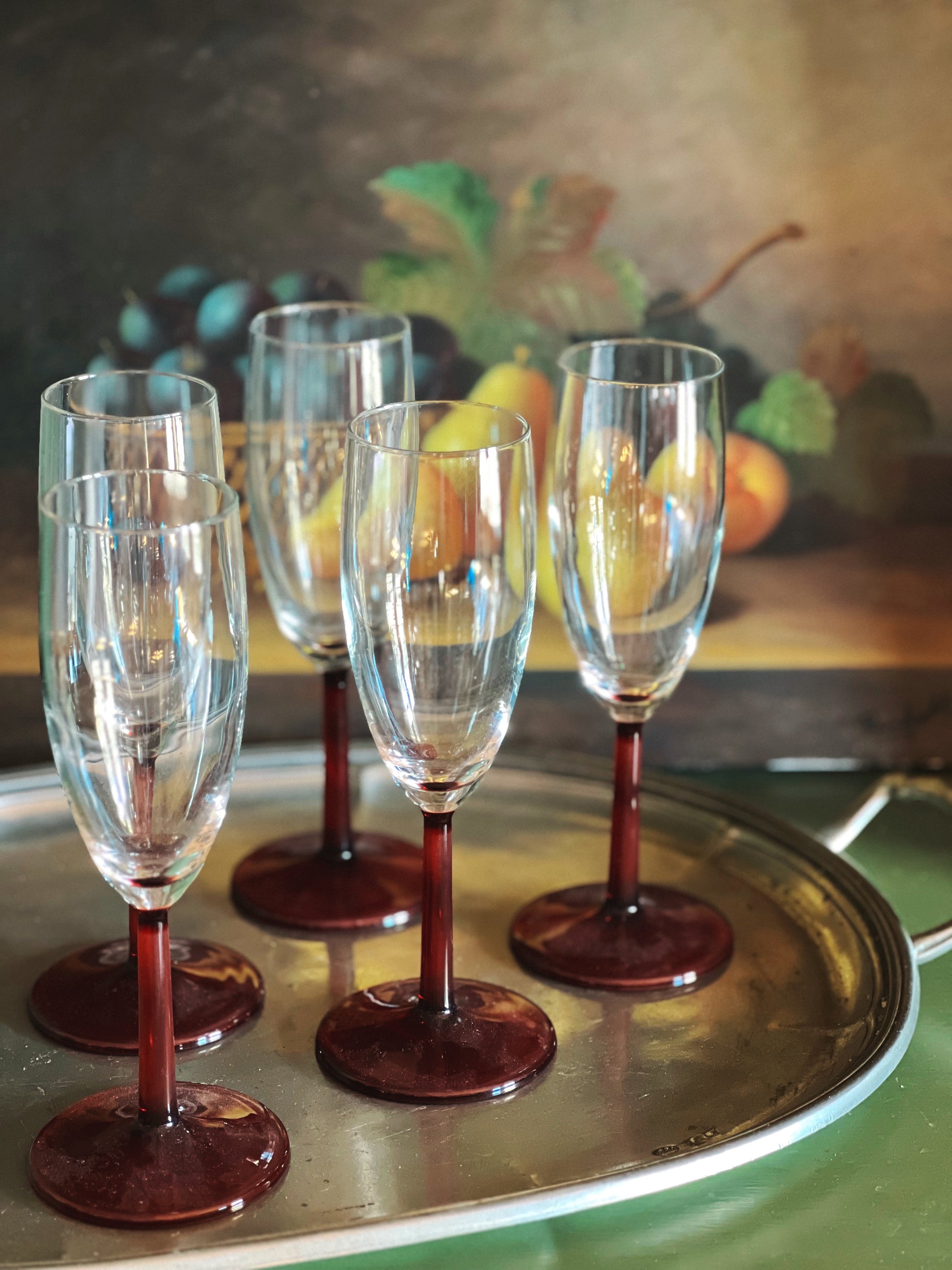 Set of champagne glasses with red bases on a gold tray in front of a painting of fruit.