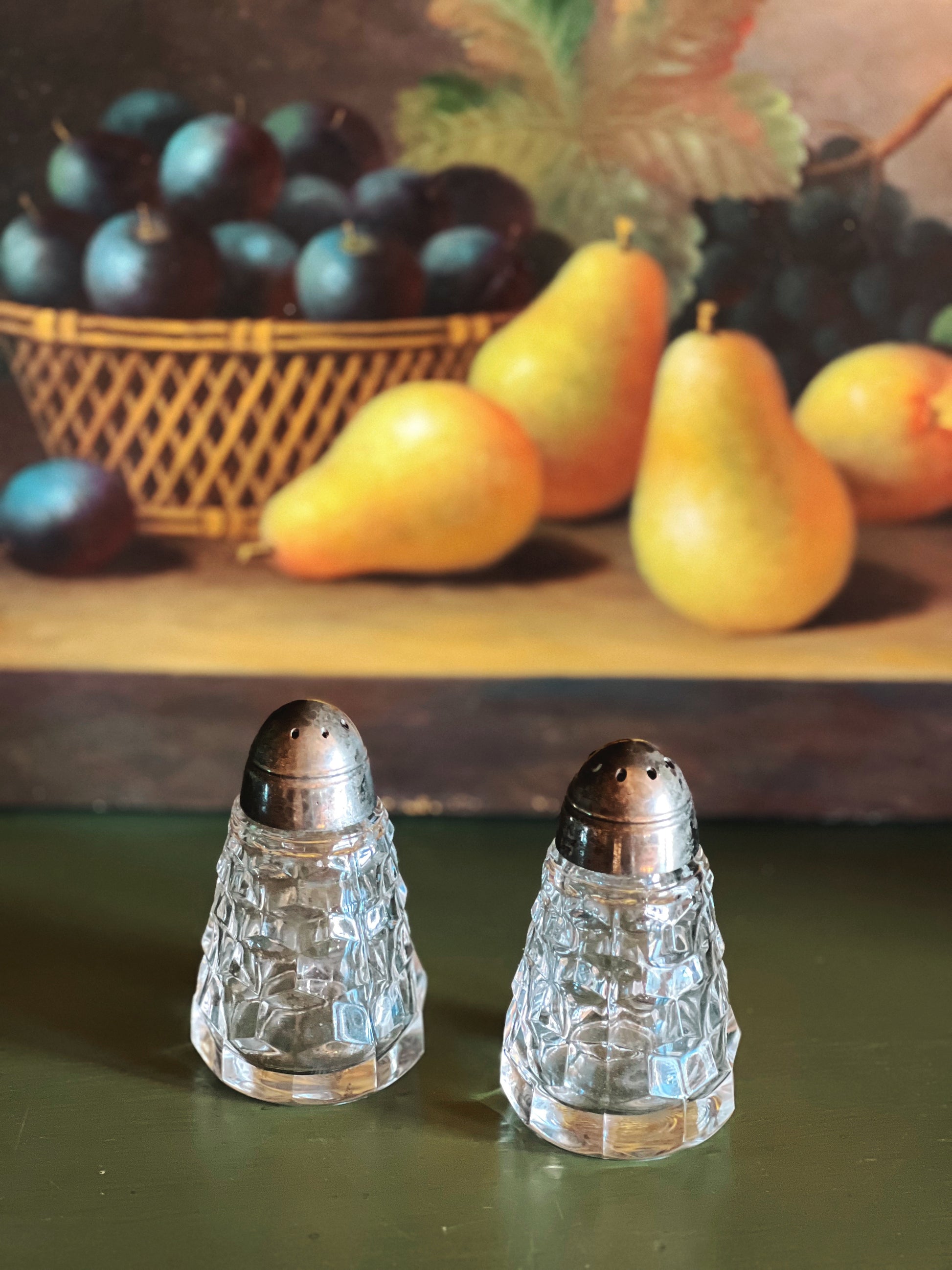 Two crystal salt shakers on a green surface with a fruit basket in the background.