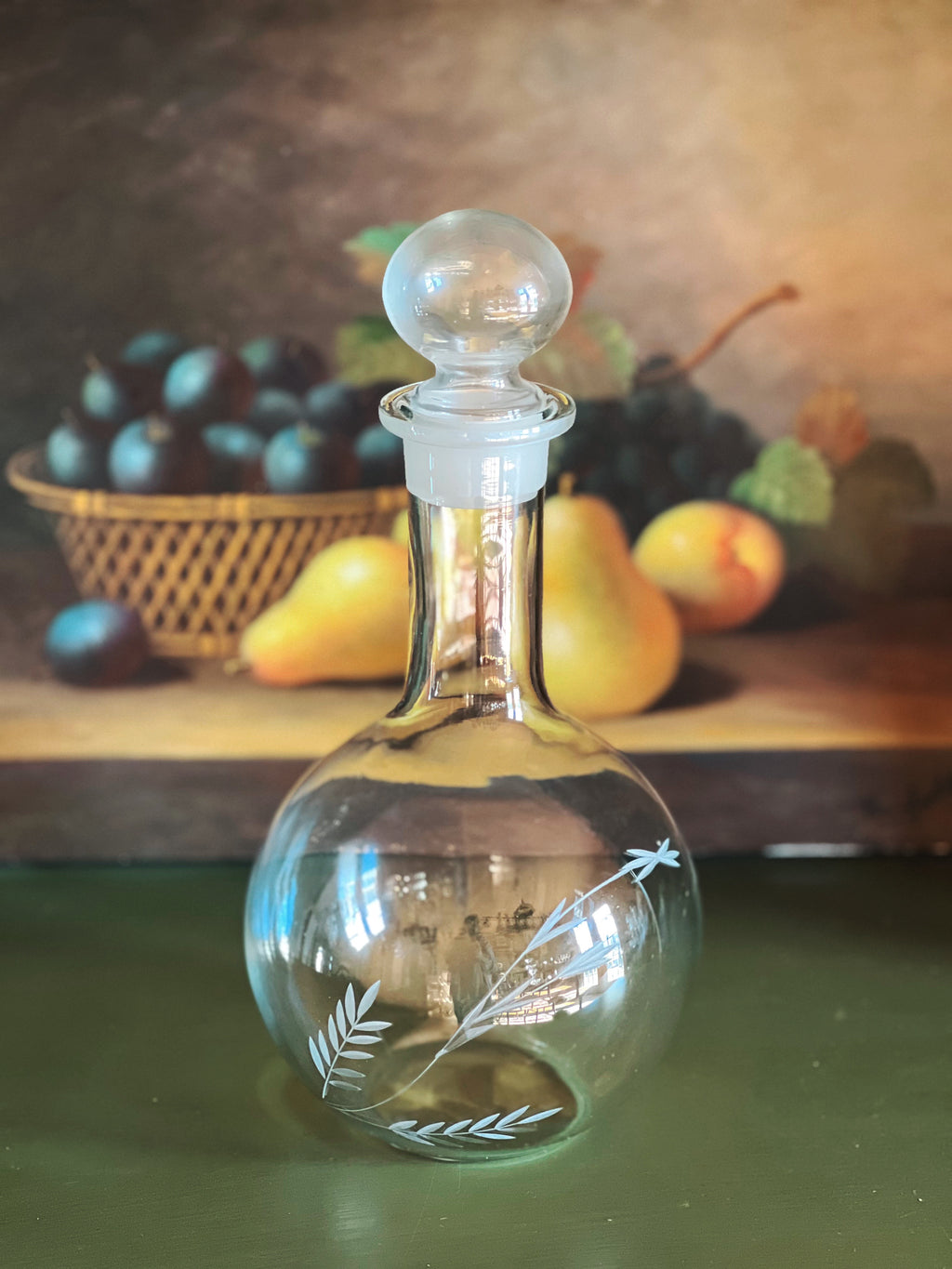 Glass decanter with etched design on a table with fruits in the background