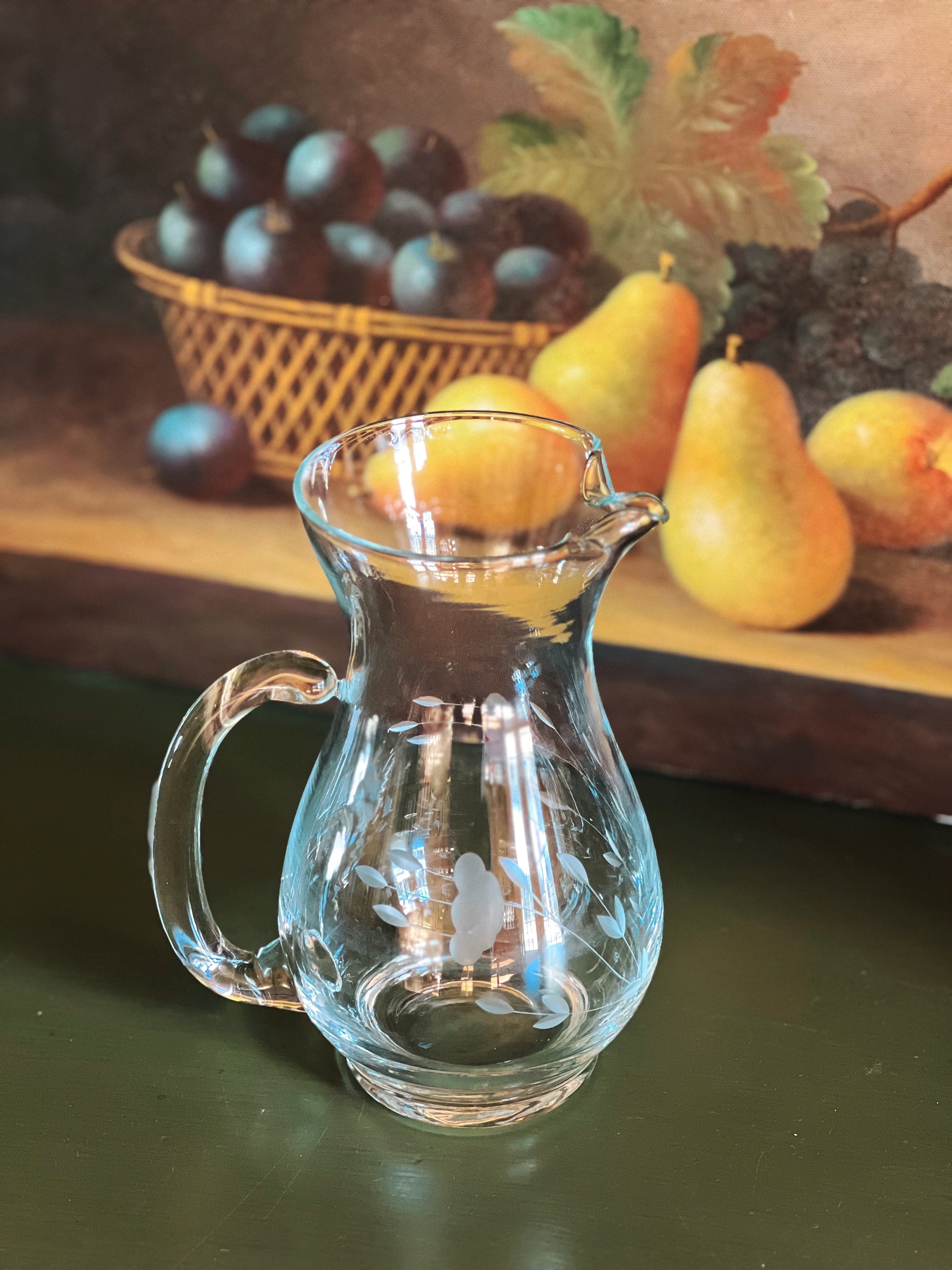 Clear glass pitcher on a green surface with fruit in the background