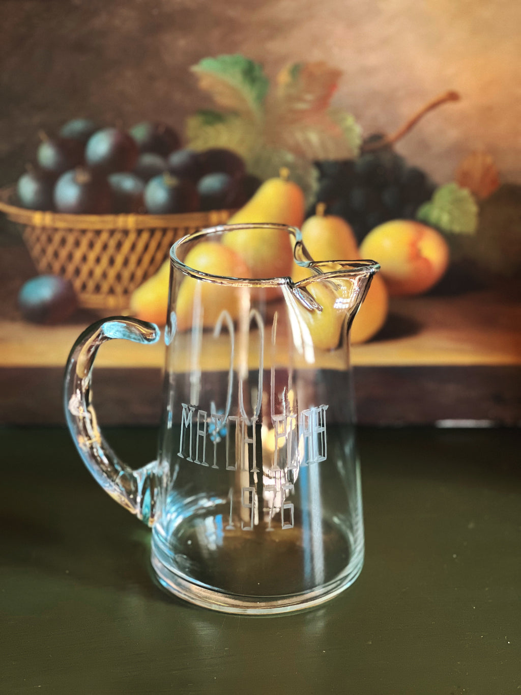 Clear glass pitcher with engraved design on a wooden surface with fruits in the background