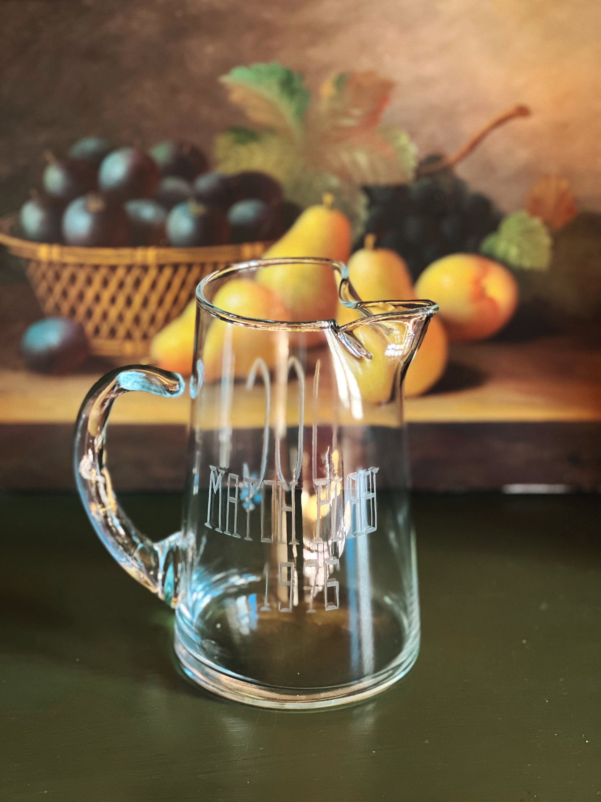 Clear glass pitcher with engraved design on a wooden surface with fruits in the background