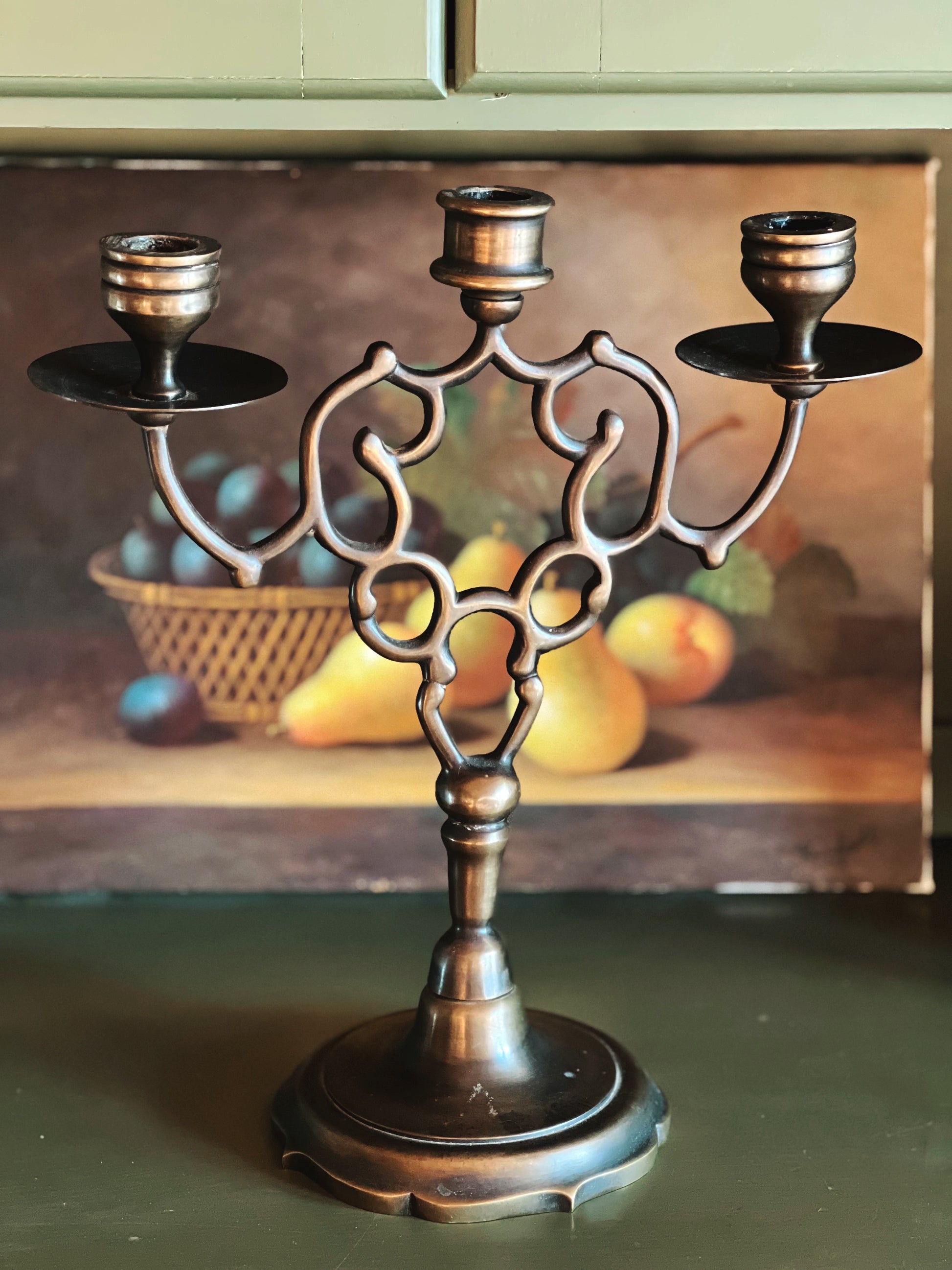 Bronze candelabra with three candle holders on a green surface, with a painting of fruit in the background.