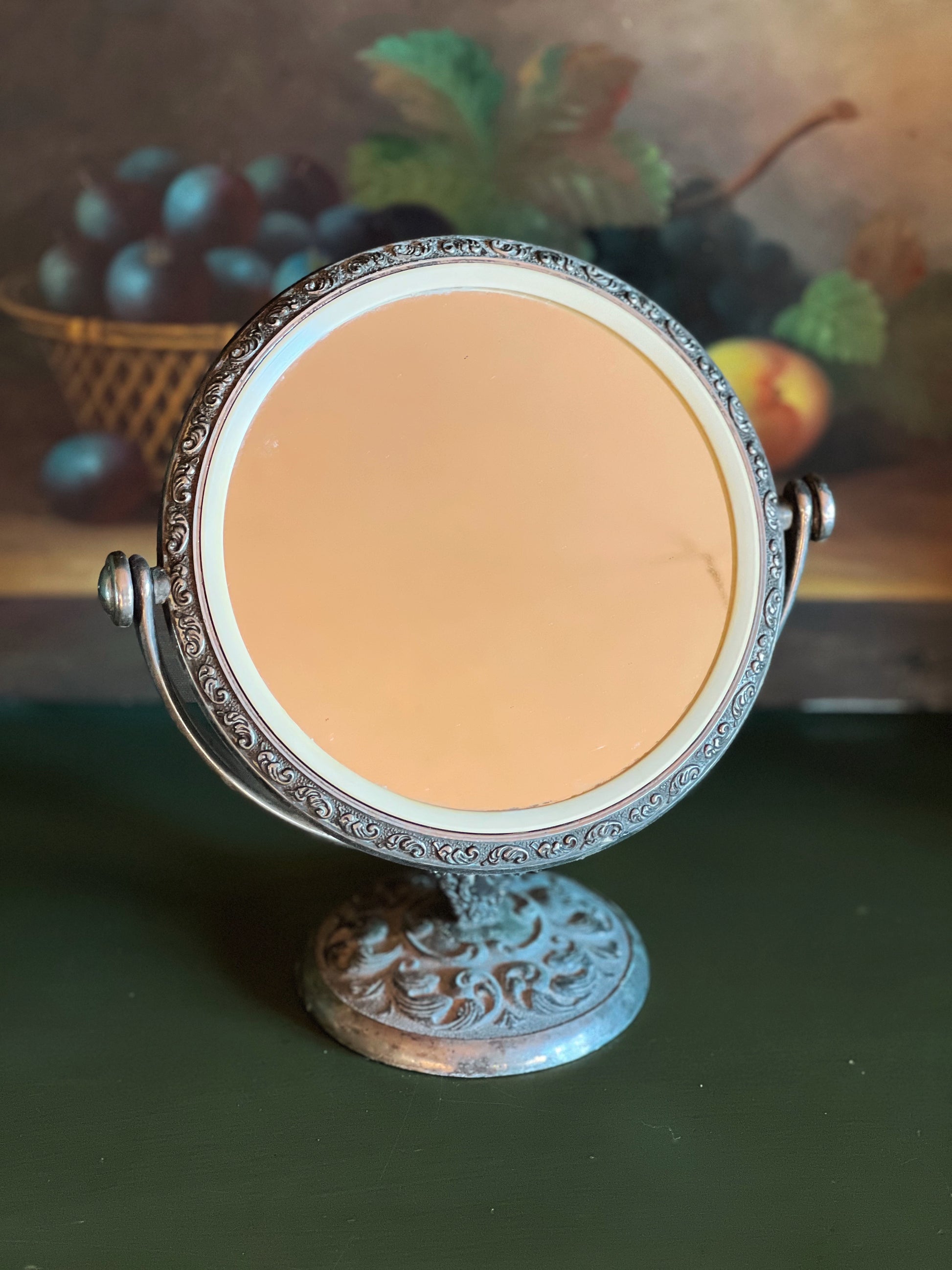 Decorative round mirror with intricate design on a stand, blurred fruit background