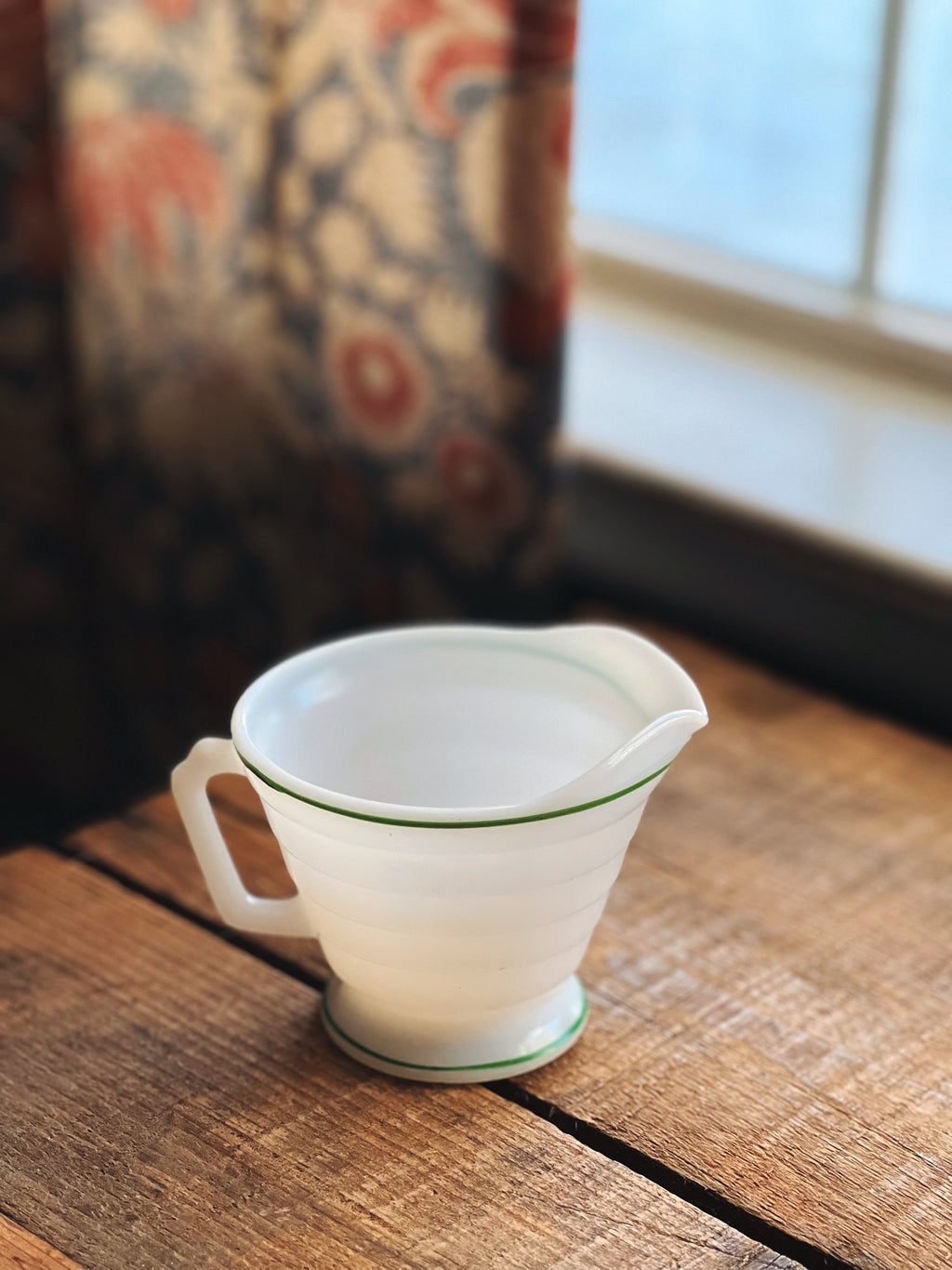 White glass measuring cup with green rim on a wooden surface near a window with patterned curtains.