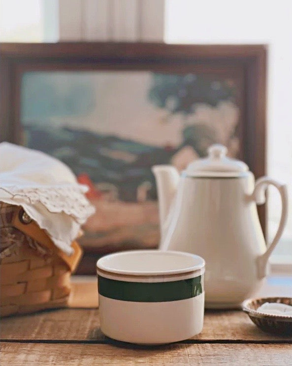 White teapot and cup with green band on a wooden surface, blurred painting in the background