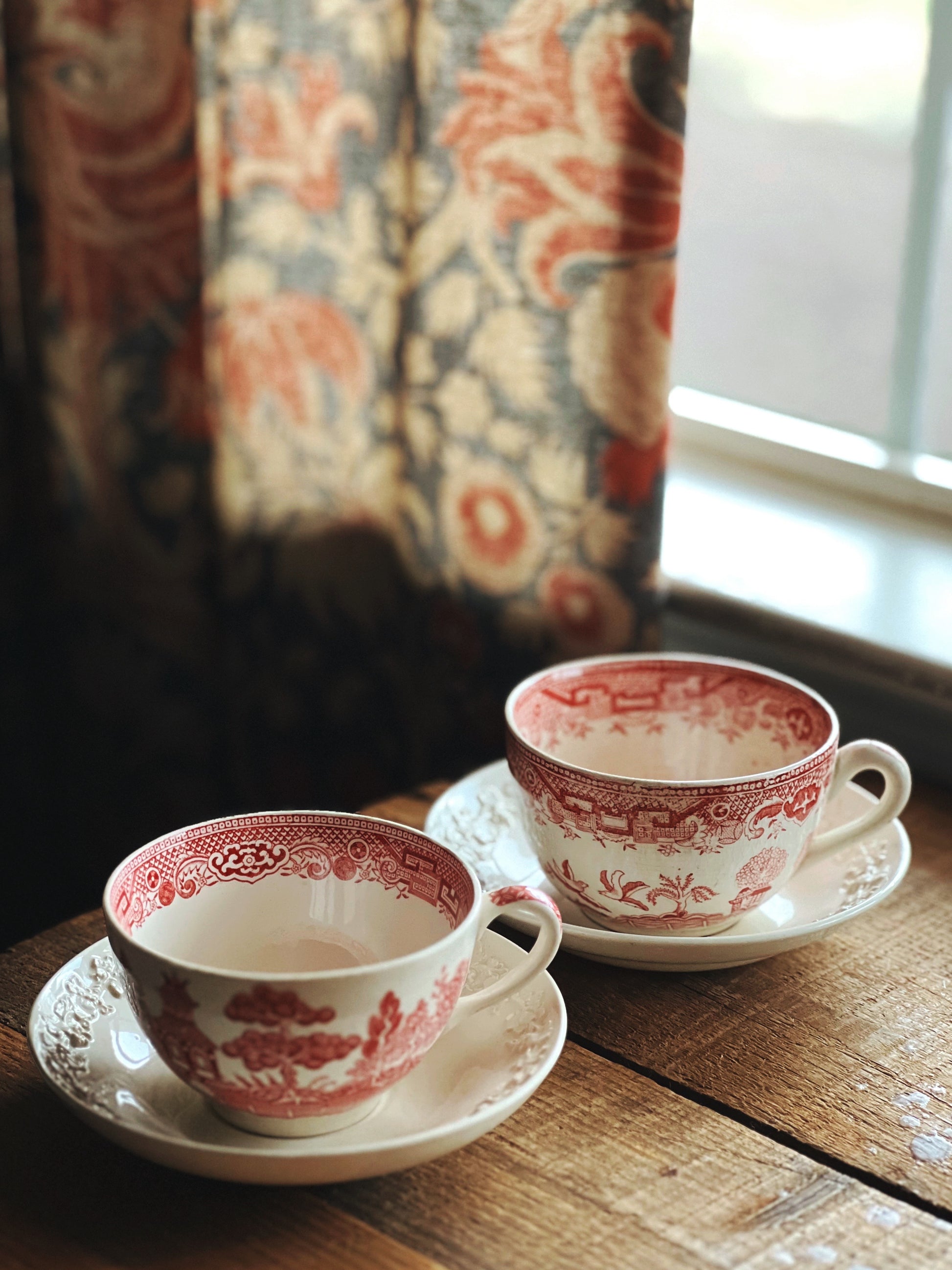 Two red and white teacups on saucers on a wooden table with a patterned curtain in the background.