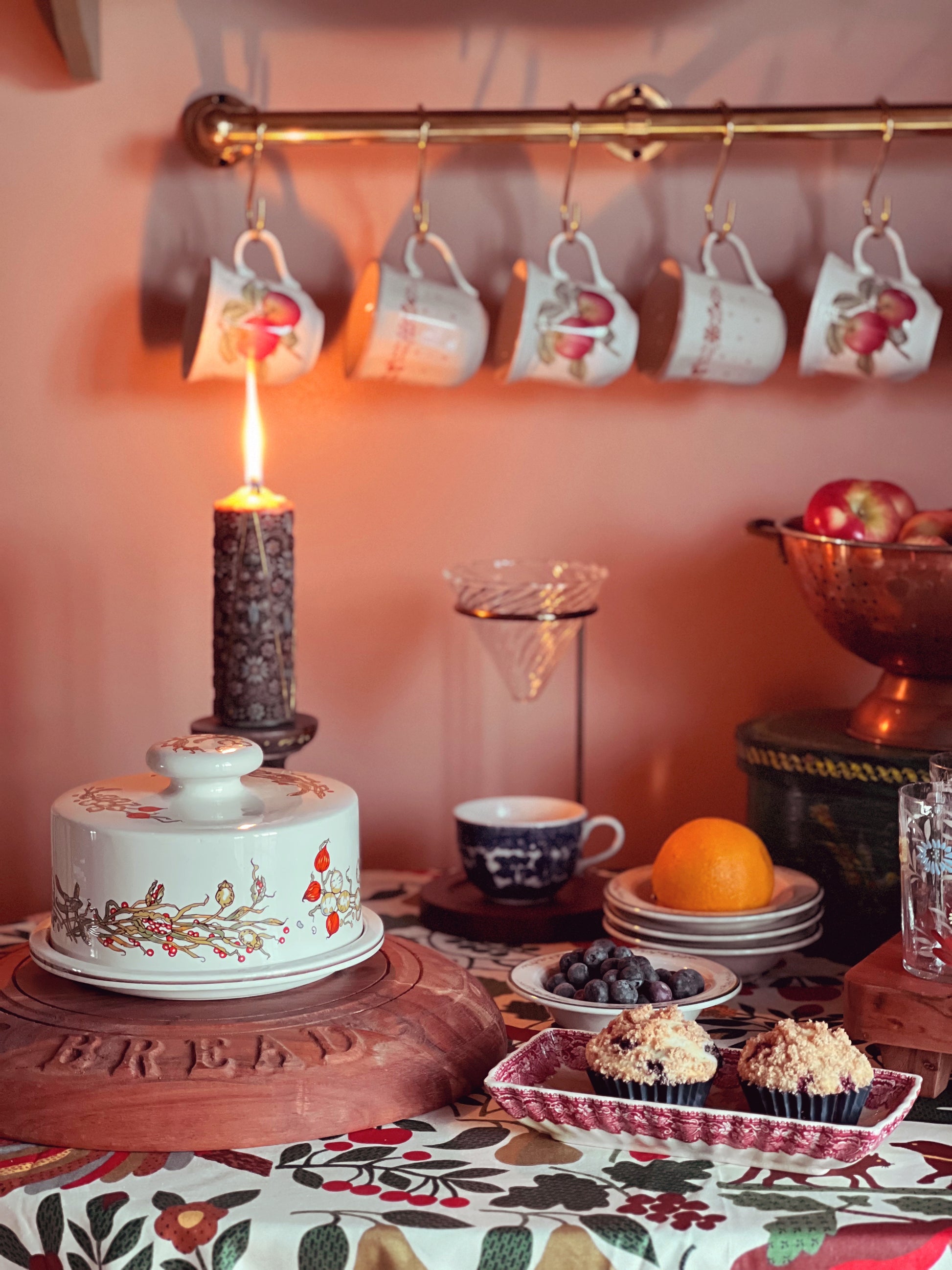 Decorative table setting with lit candle, cake, and fruit on a floral tablecloth.