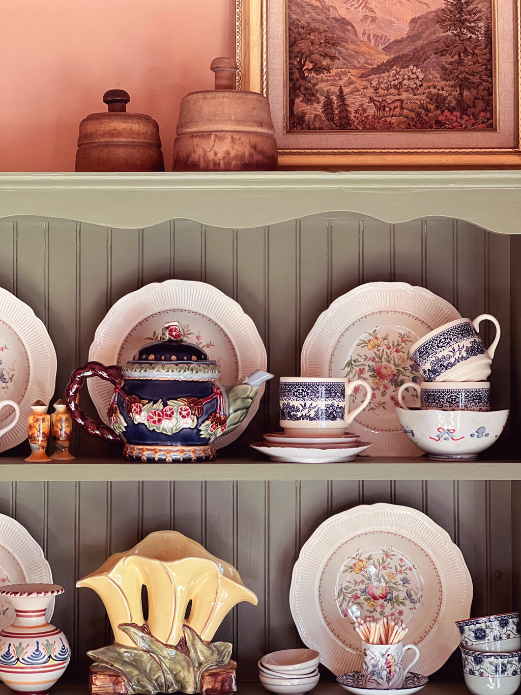 Collection of decorative ceramic items on a shelf against a wall with a framed landscape painting.