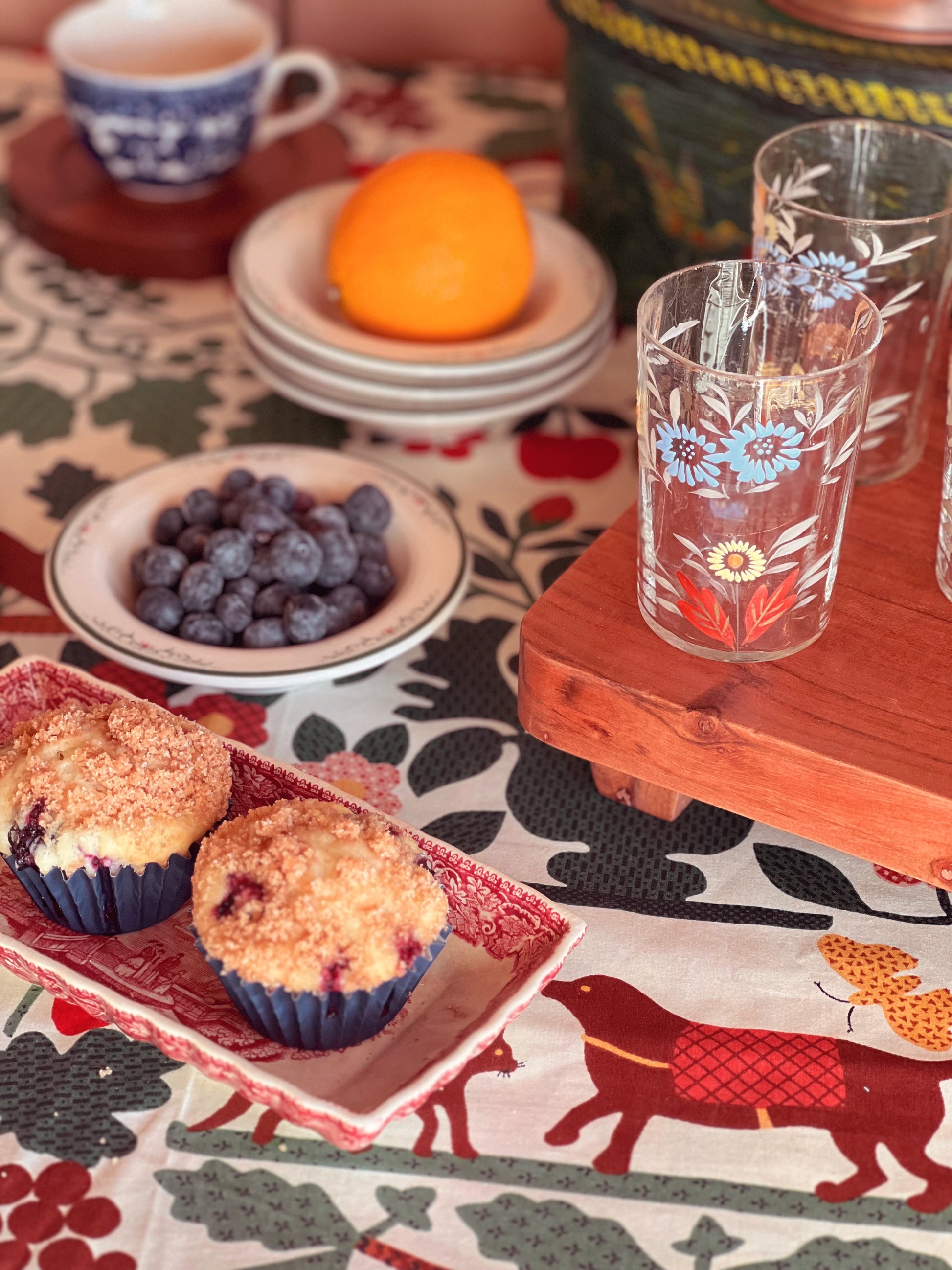 Muffins on a tray with a bowl of blueberries, an orange, and floral-patterned glasses on a patterned tablecloth.