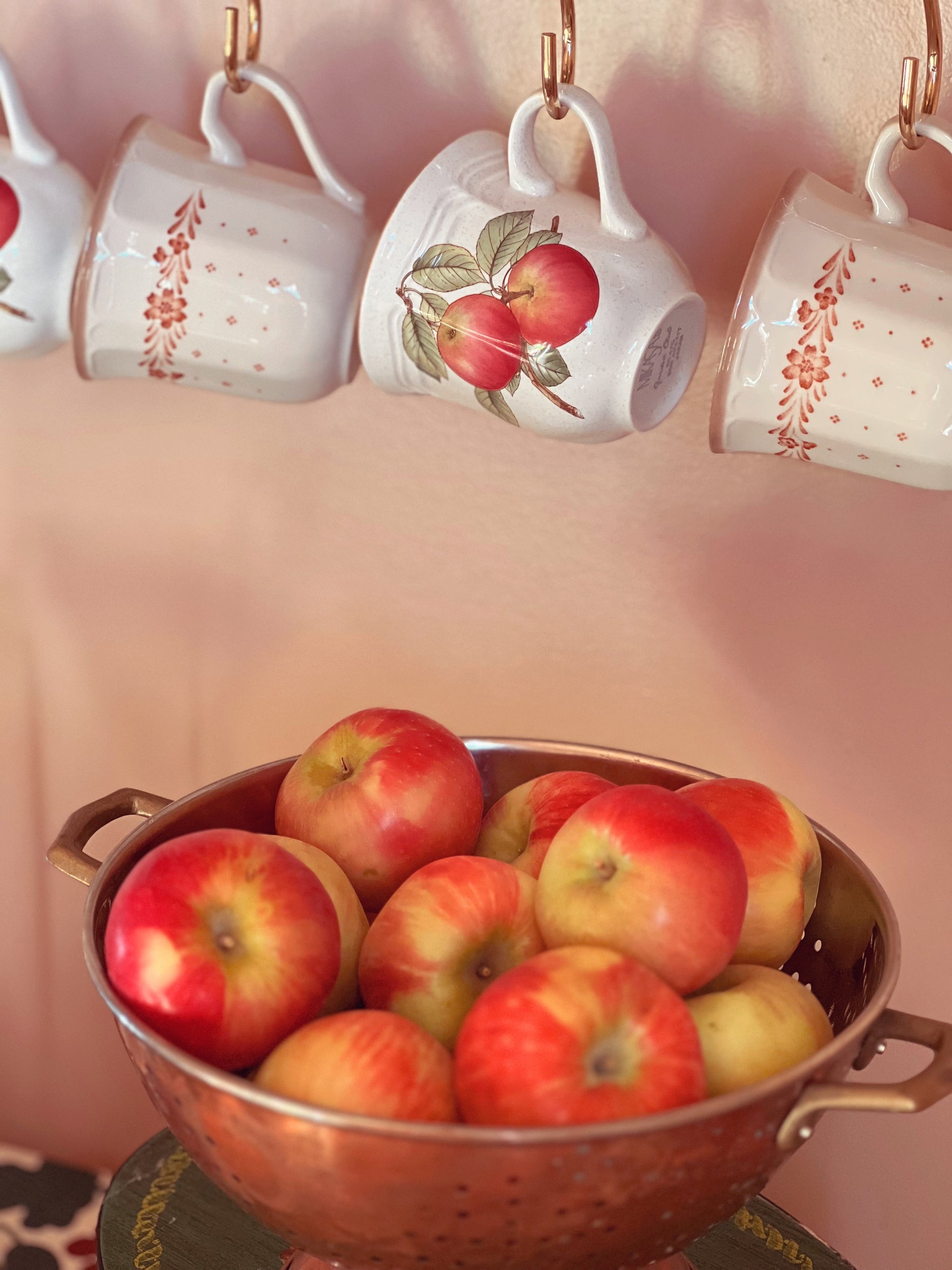 Decorative mugs with fruit designs hanging above a colander filled with apples.