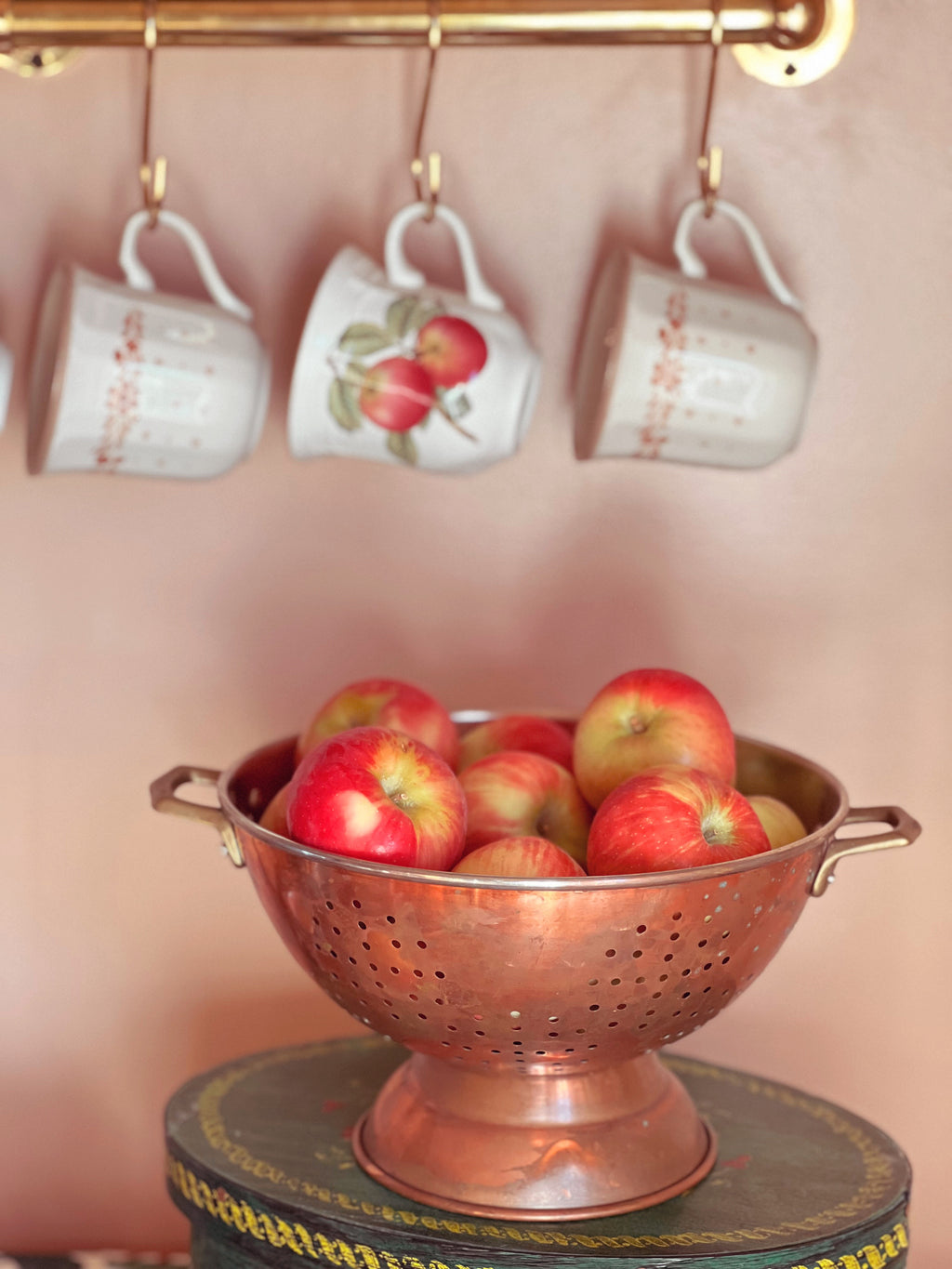 Copper colander filled with apples on a stand with hanging mugs in the background.