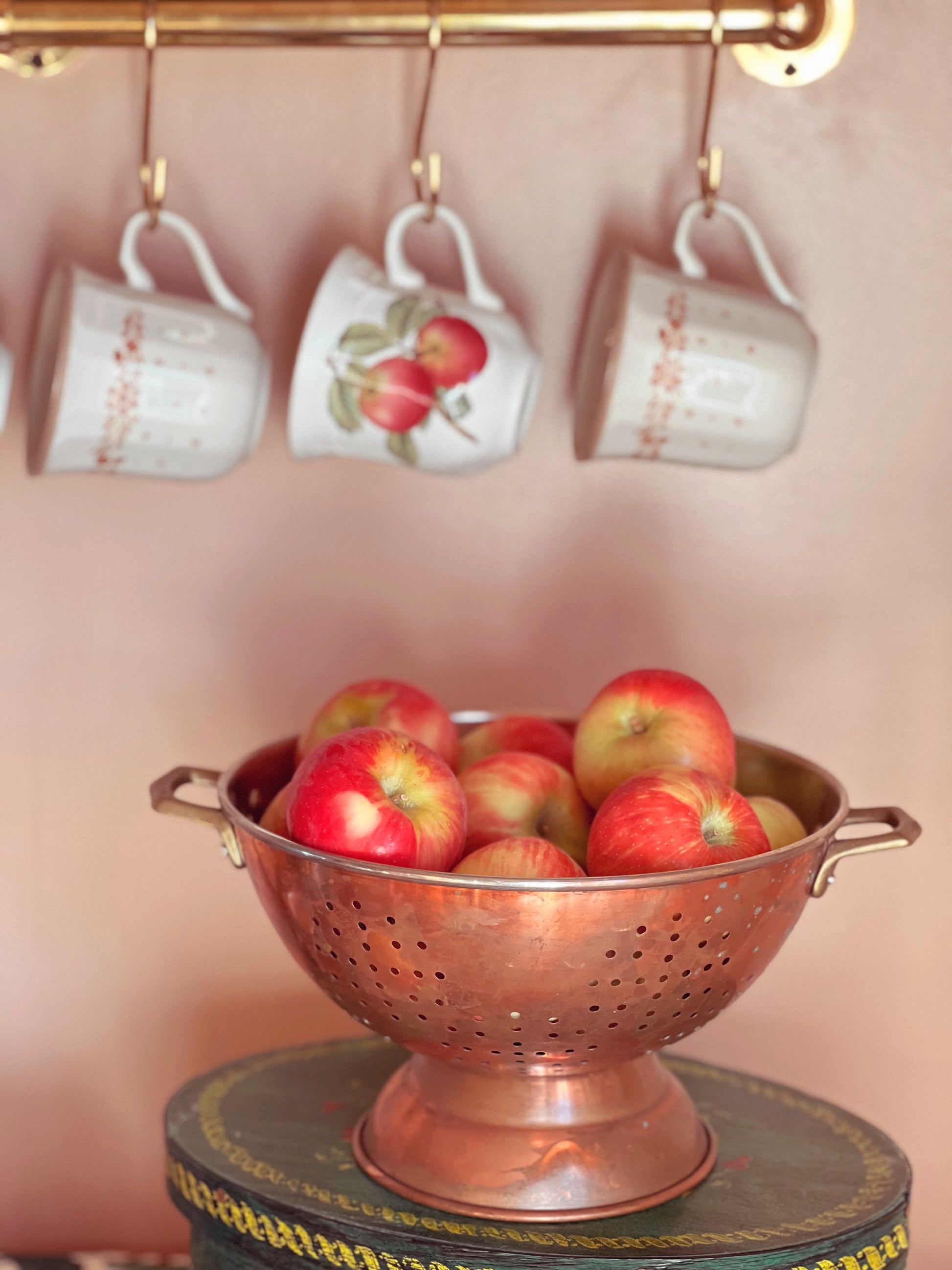 Copper colander filled with apples on a stand with hanging mugs in the background.