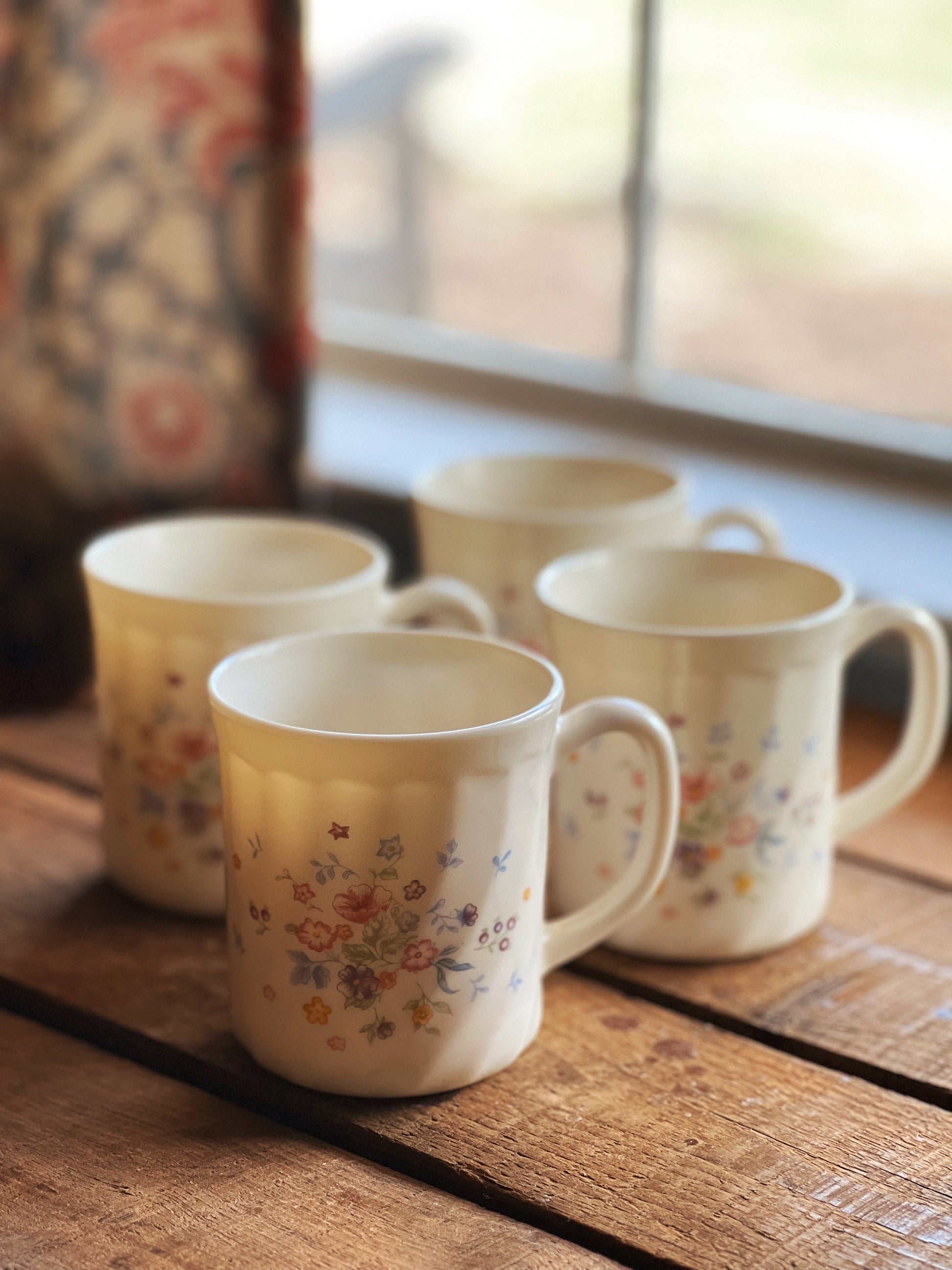 Four ceramic mugs with floral patterns on a wooden surface near a window.