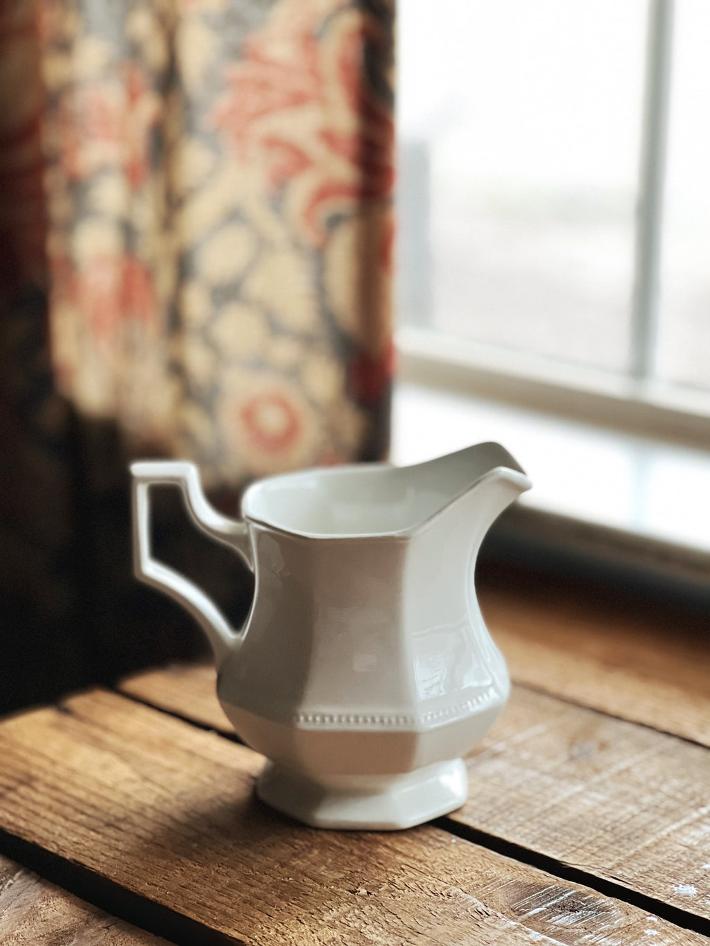 White ceramic pitcher on a wooden surface with floral curtains in the background