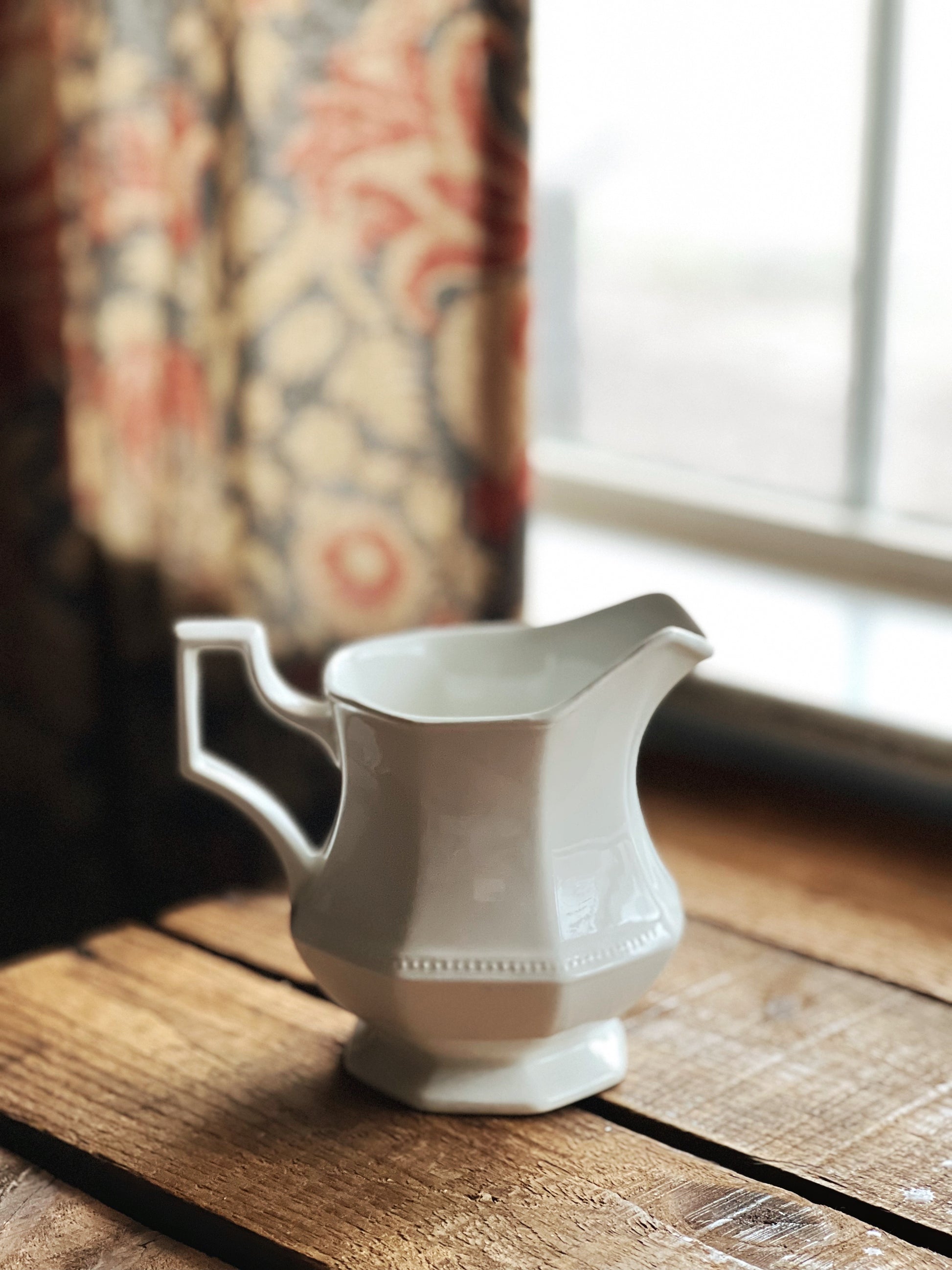 White ceramic pitcher on a wooden surface with floral curtains in the background