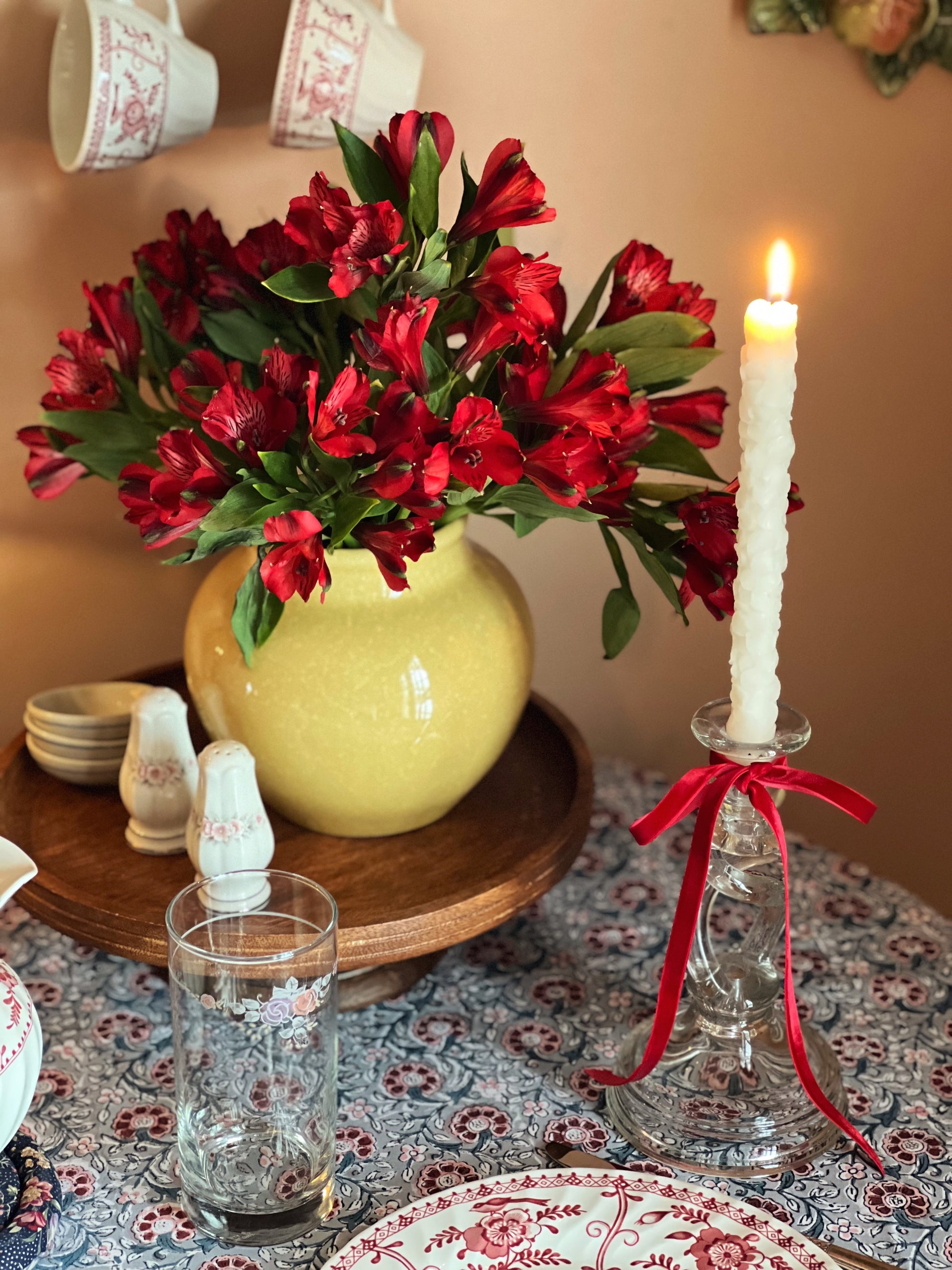 Decorative table setting with red flowers in a yellow vase, a lit candle, and glassware on a patterned tablecloth.