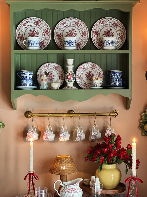Decorative shelf with floral plates, hanging teacups, and a vase of red flowers.