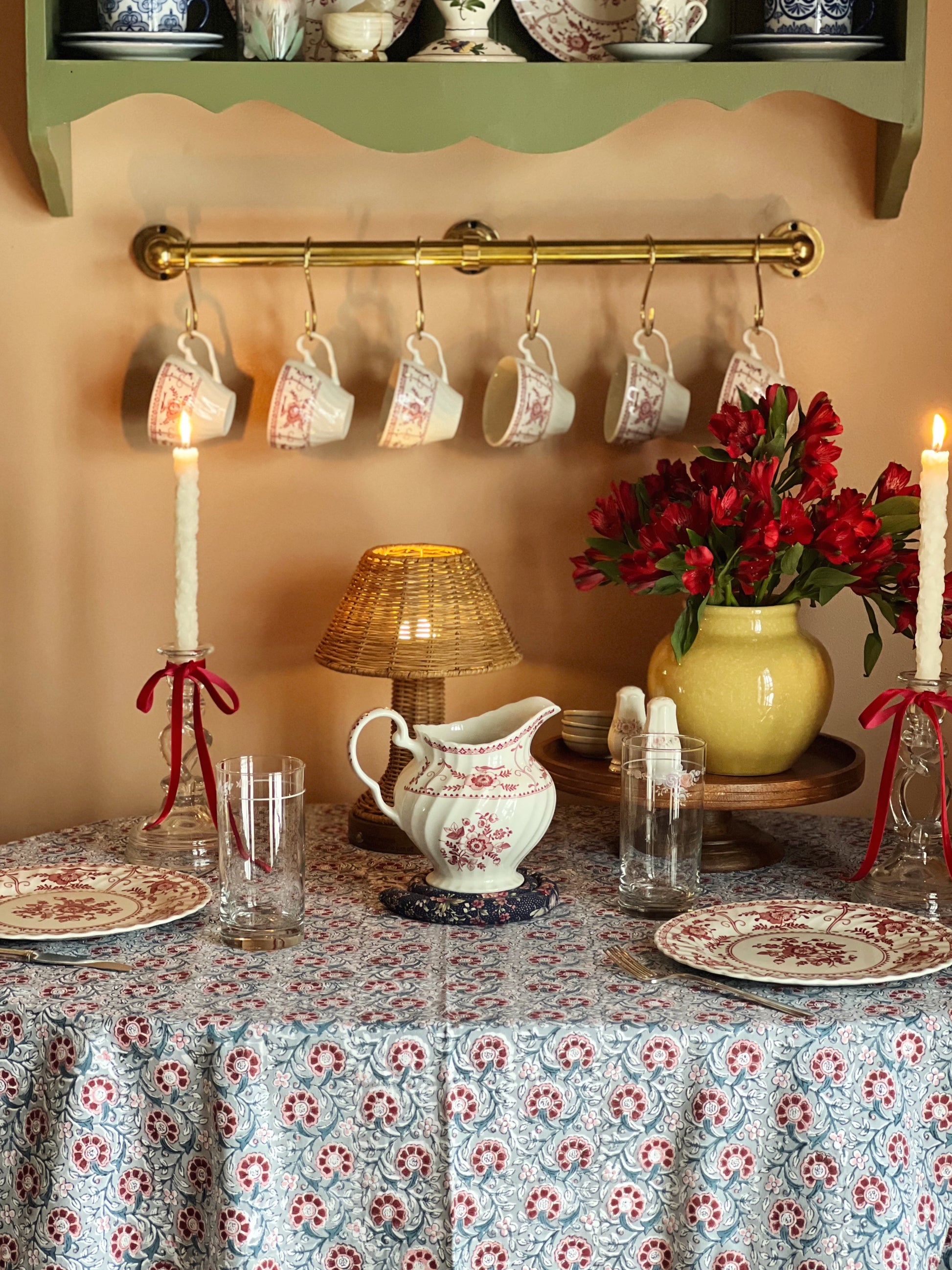 Decorative table setting with floral tablecloth, candles, and teacups on a shelf.