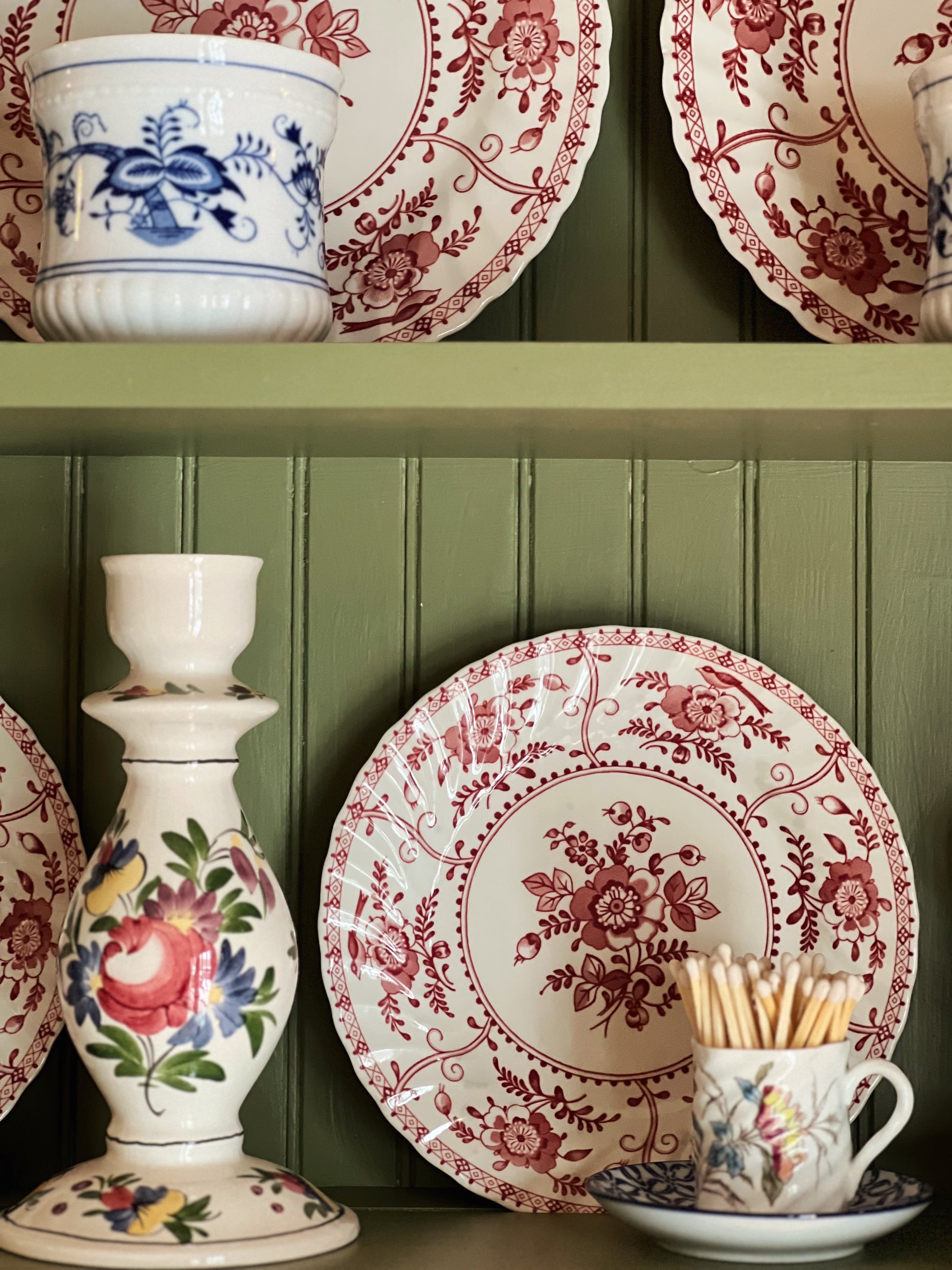 Decorative ceramic items including plates, a candlestick, and a cup with saucer on a green paneled wall background.