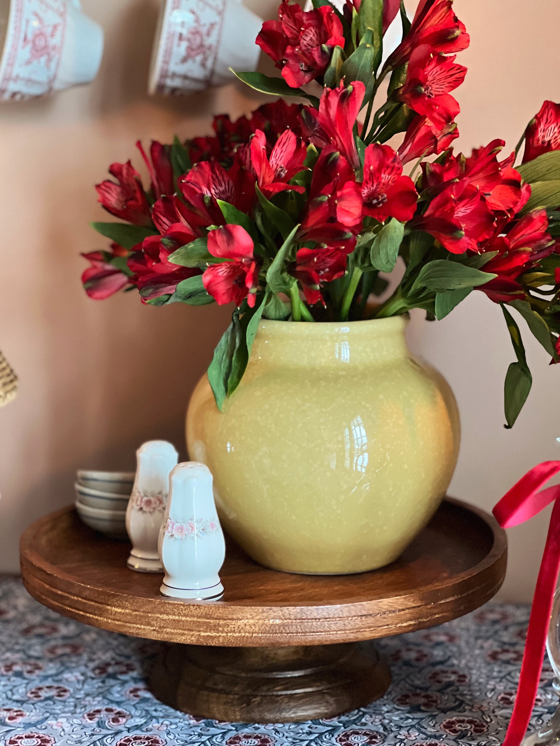 Green vase with red flowers on a wooden stand against a pink wall.