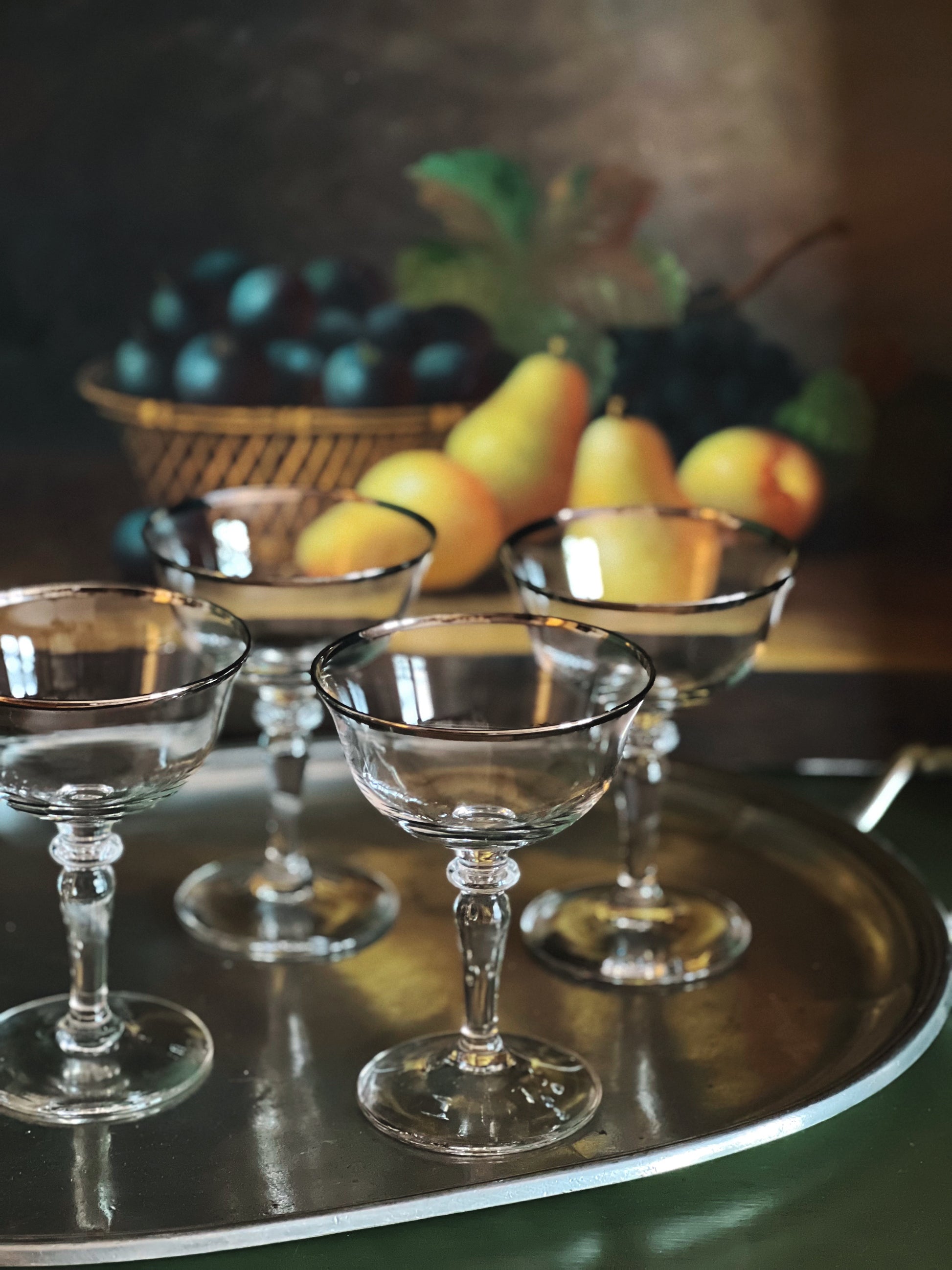 Clear glass goblets on a reflective surface with fruit in the background