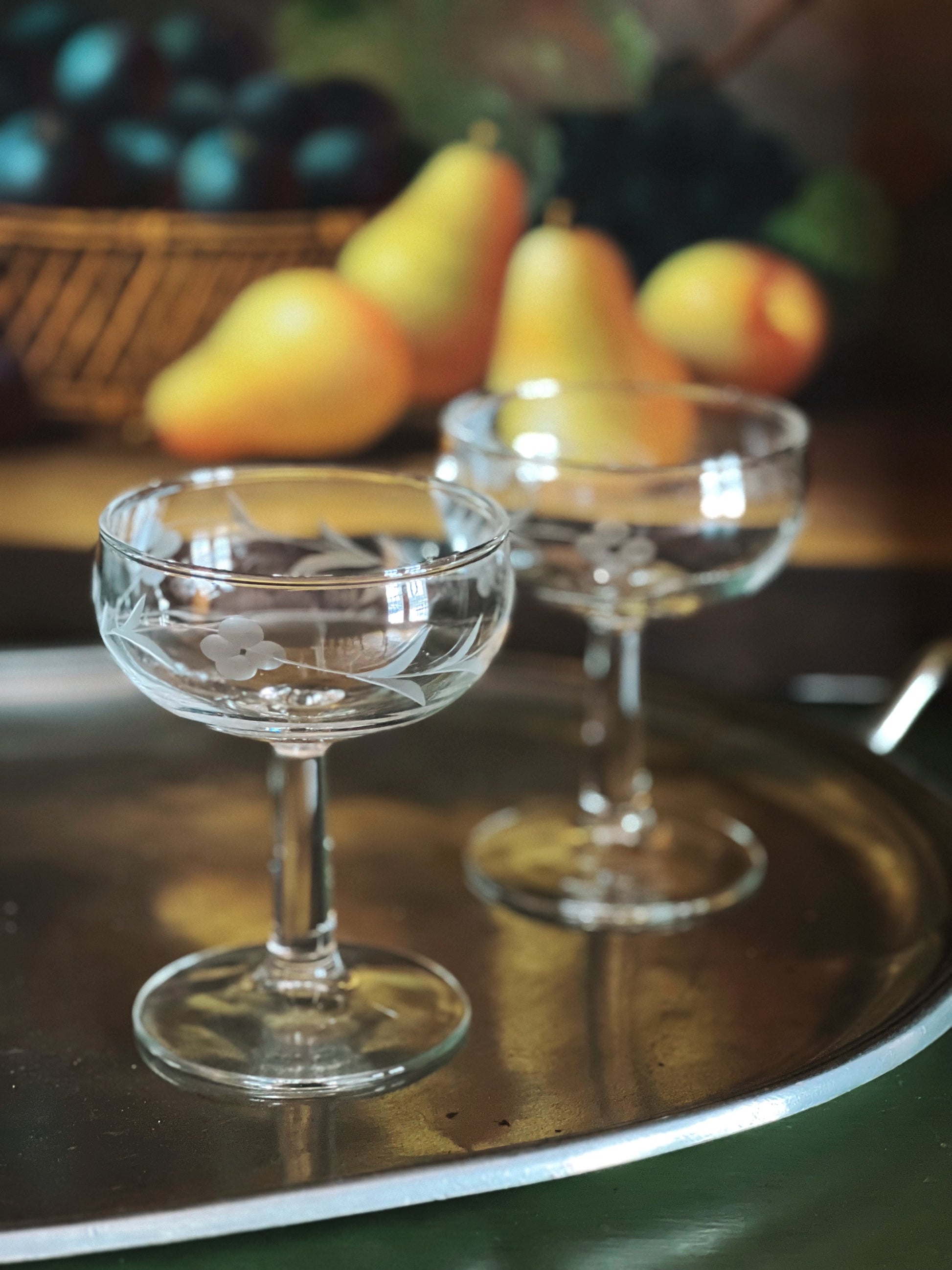 Two clear glass coupes on a reflective surface with blurred fruit in the background