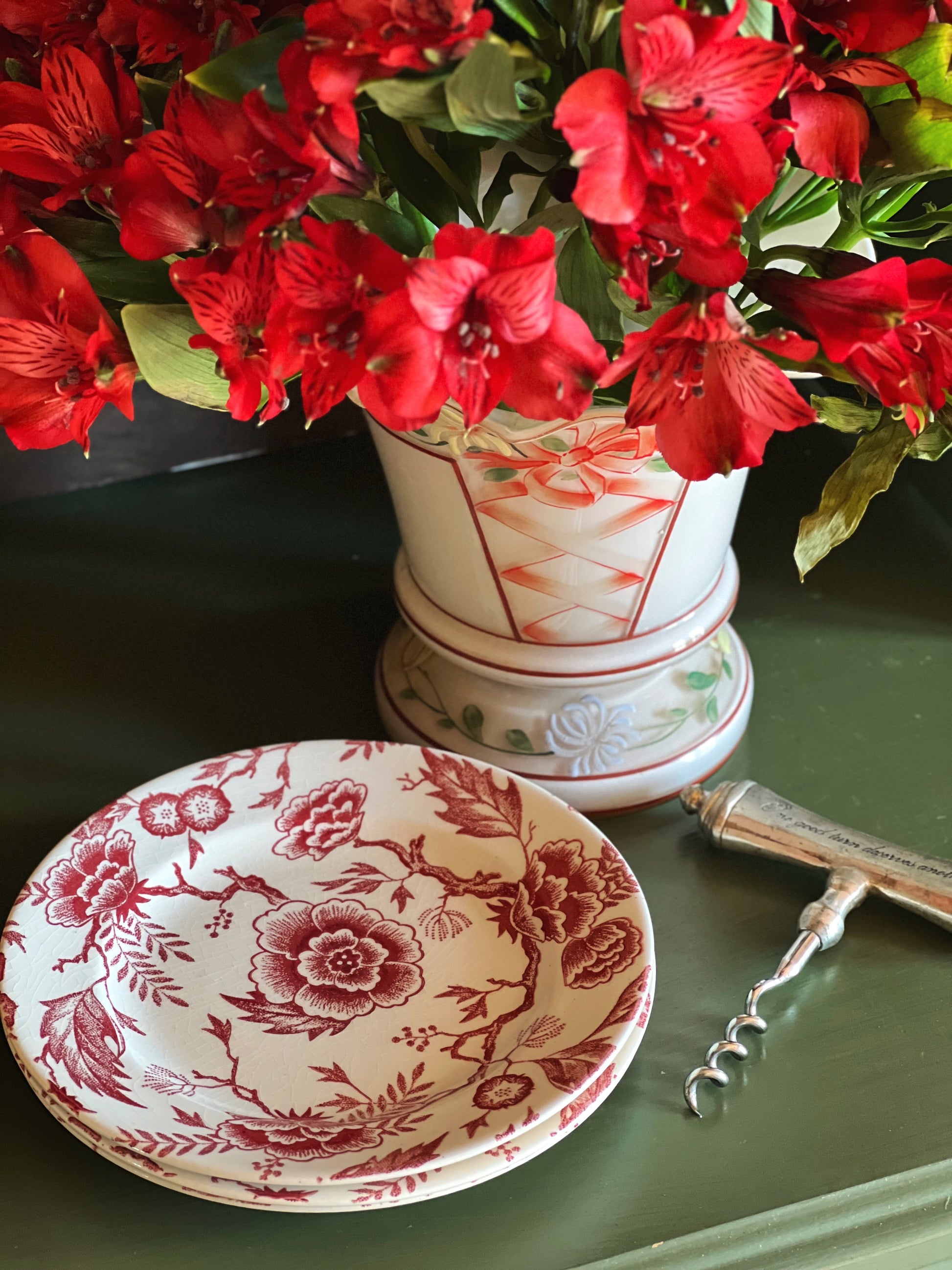 Decorative vase with red flowers on a green surface, accompanied by a matching plate and corkscrew.