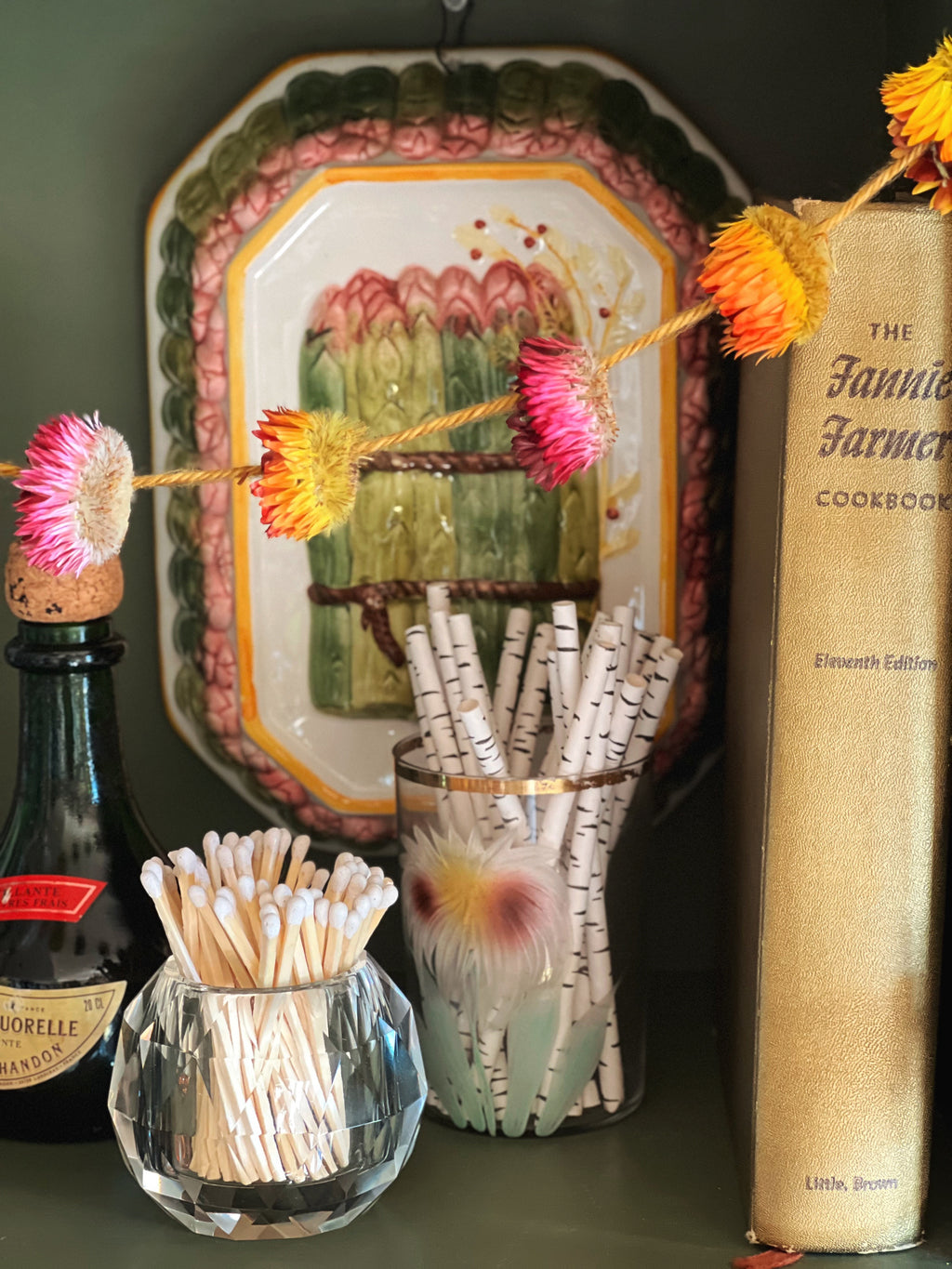 Decorative setting with a bottle, vintage cookbook, and decorative items on a dark surface.