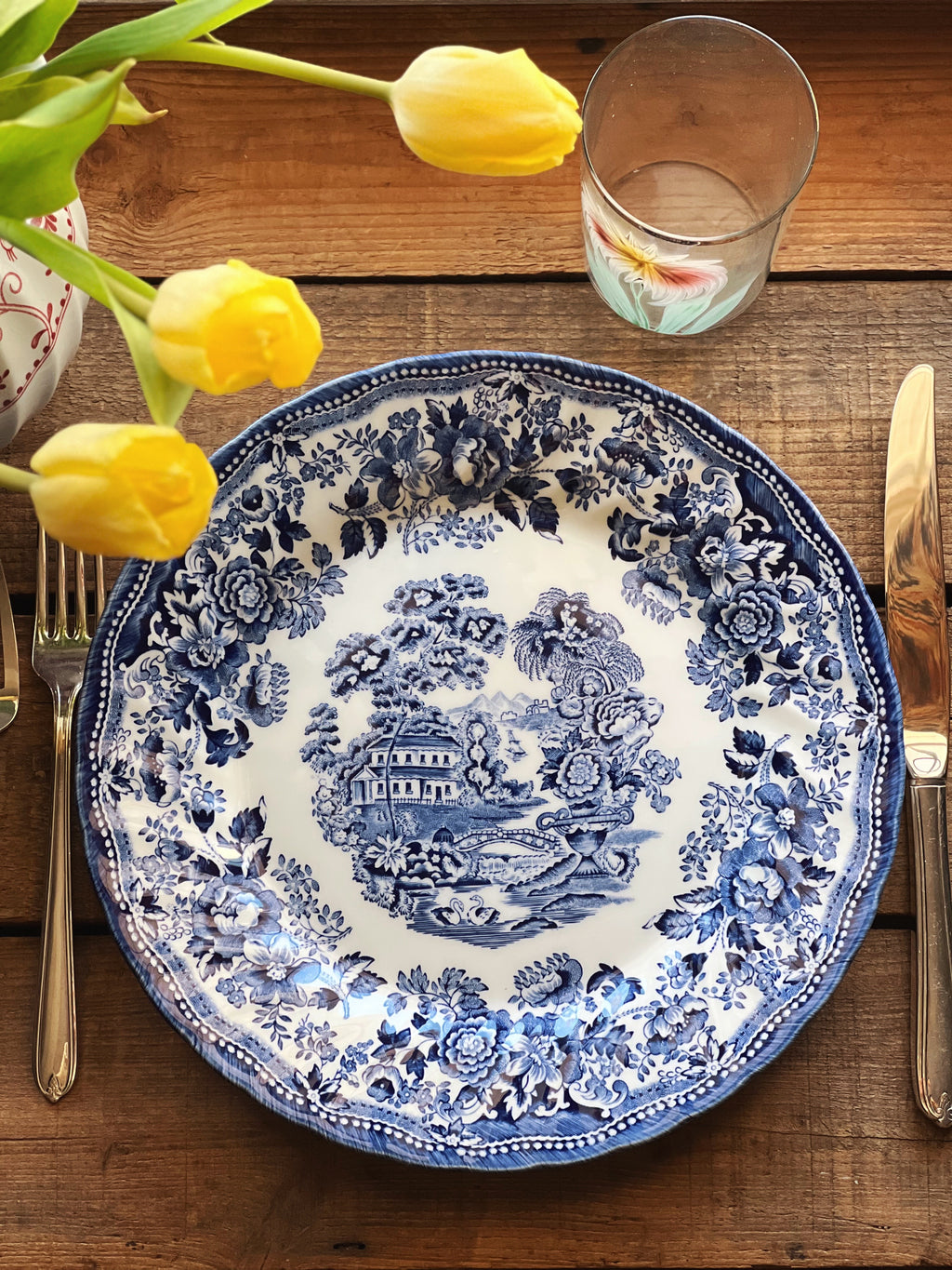 Decorative blue and white plate on a wooden table with yellow tulips and cutlery.