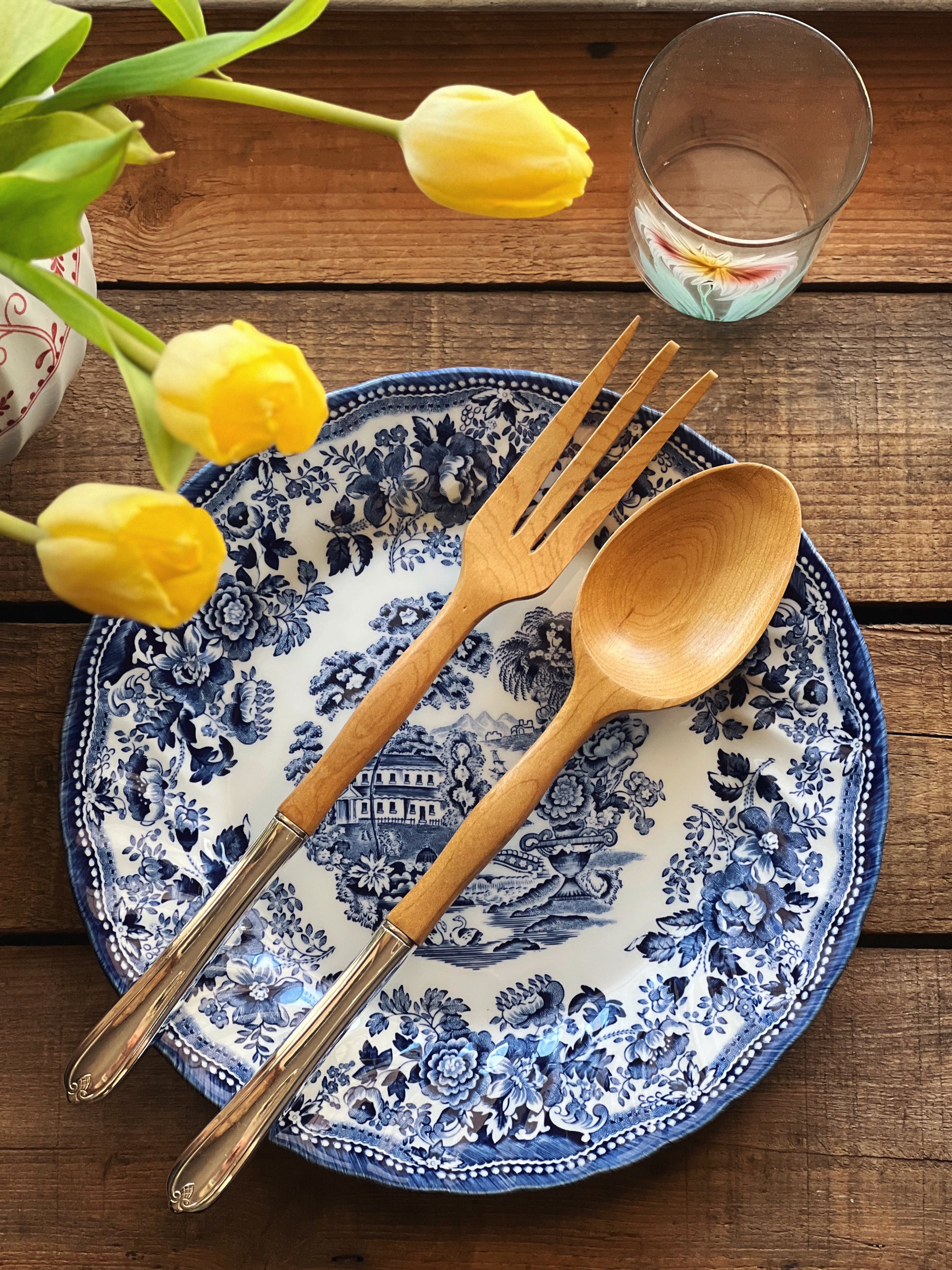 Wooden fork and spoon on a blue and white floral plate with yellow tulips and a glass in the background.
